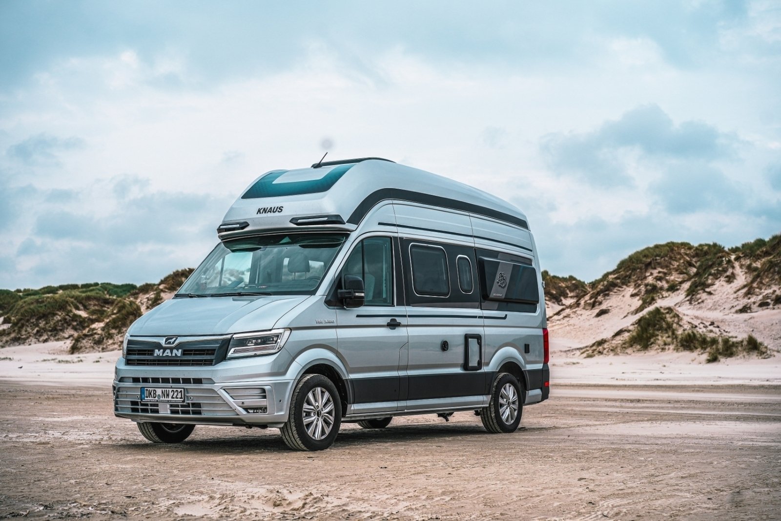 A silver Knaus van parked on a sandy beach with dunes in the background under a partly cloudy sky.