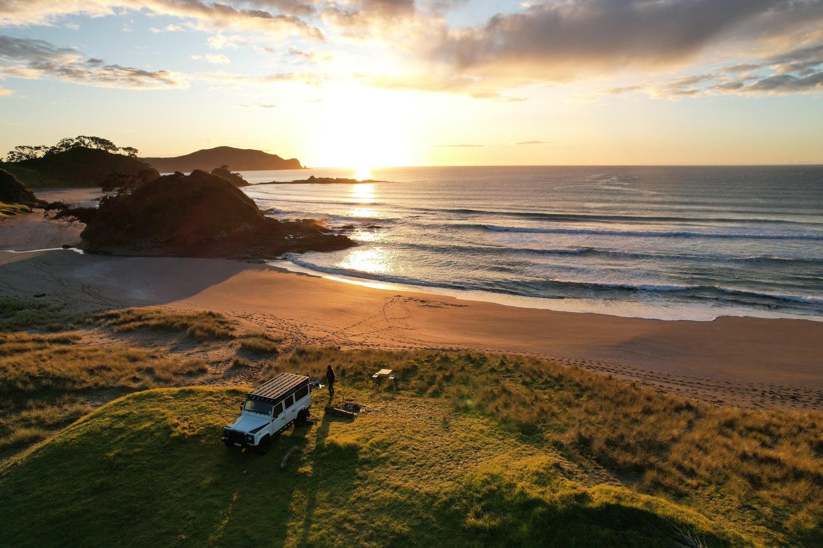 A scenic coastal view at sunrise, with a vehicle parked on grassy terrain overlooking a sandy beach and calm ocean waves.