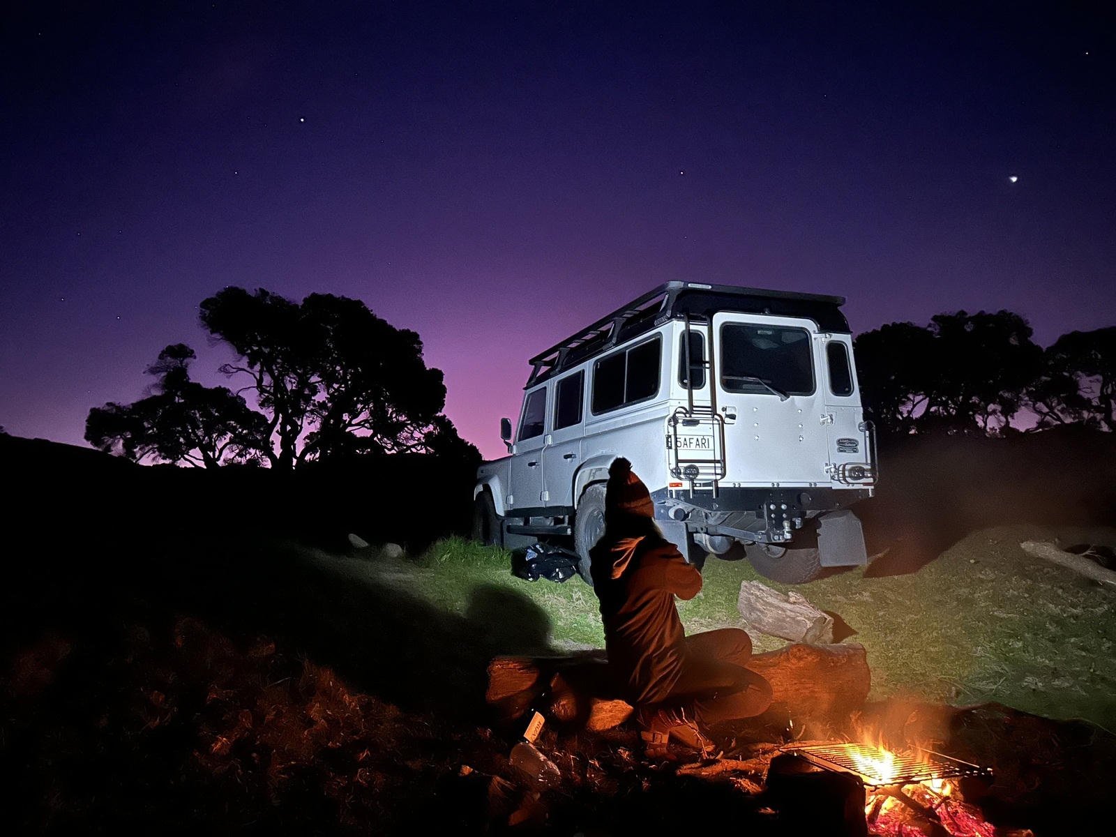 Person sitting by a campfire near a parked van under a twilight sky with visible stars and silhouetted trees.