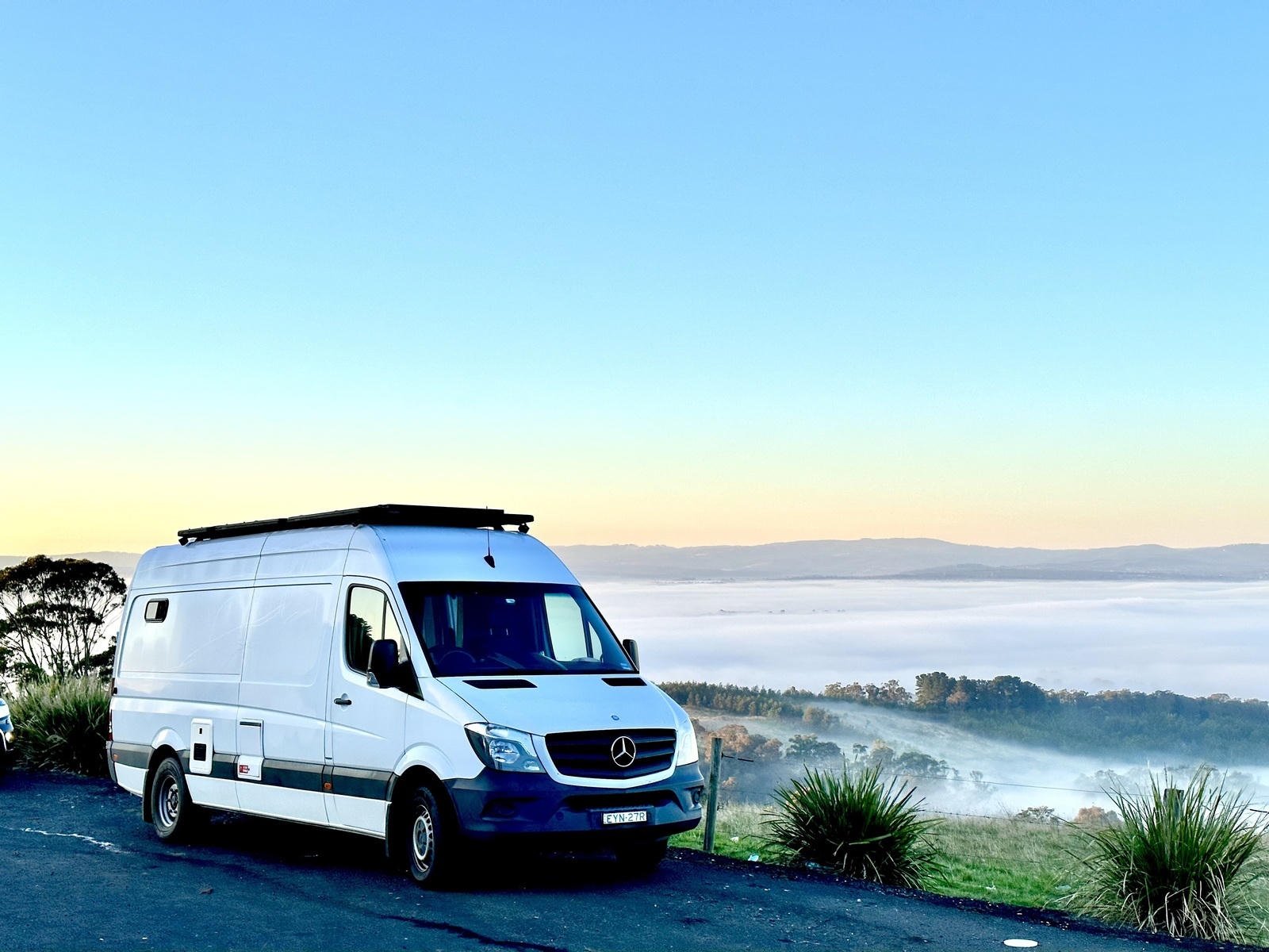 White van parked on a scenic overlook, with a misty valley and distant hills under a clear blue sky.