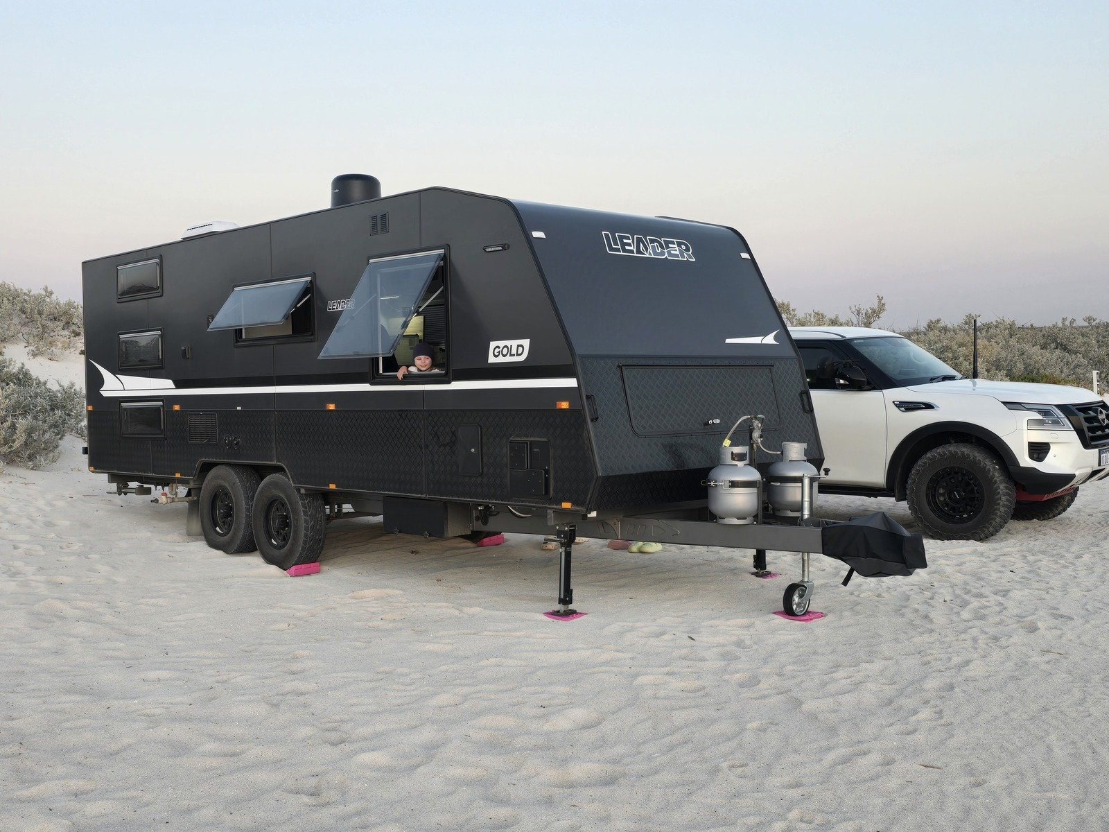 A Leader caravan hired on Camplify parked on sandy terrain beside a white SUV, featuring a child peeking through a window.
