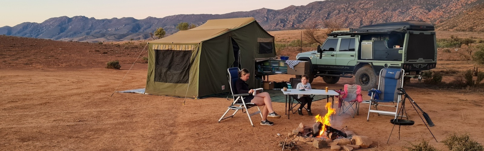 Camplify Camping scene with green tent, SUV, and people sitting around a campfire in a desert landscape with mountains in the background.