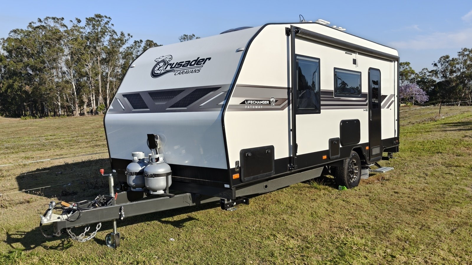 A Crusader Caravan hired on Camplify parked on grass, with trees in the background and two gas cylinders at the front.