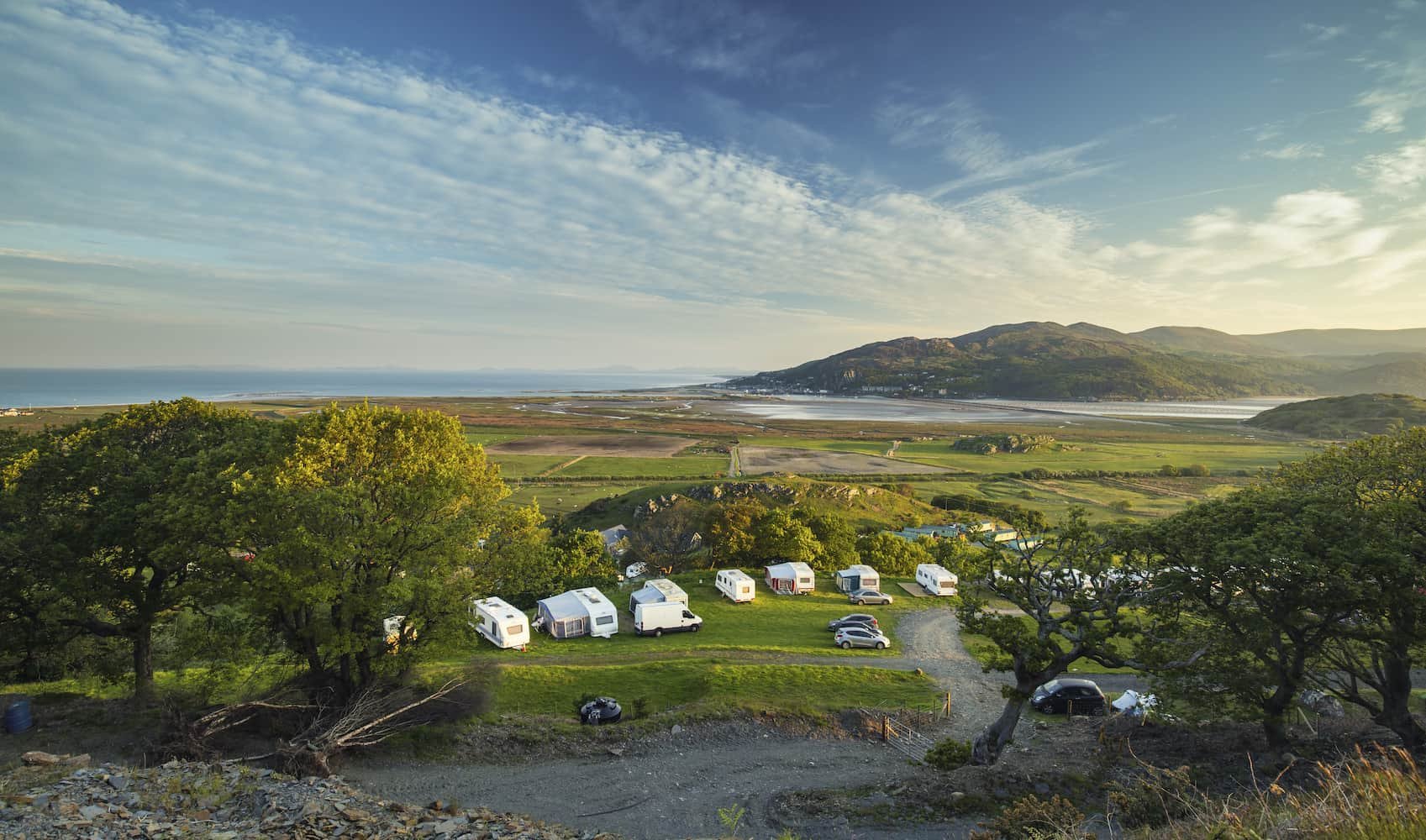 A scenic view of a campsite with several caravans, surrounded by trees, overlooking a coastal landscape and distant mountains under a blue sky.