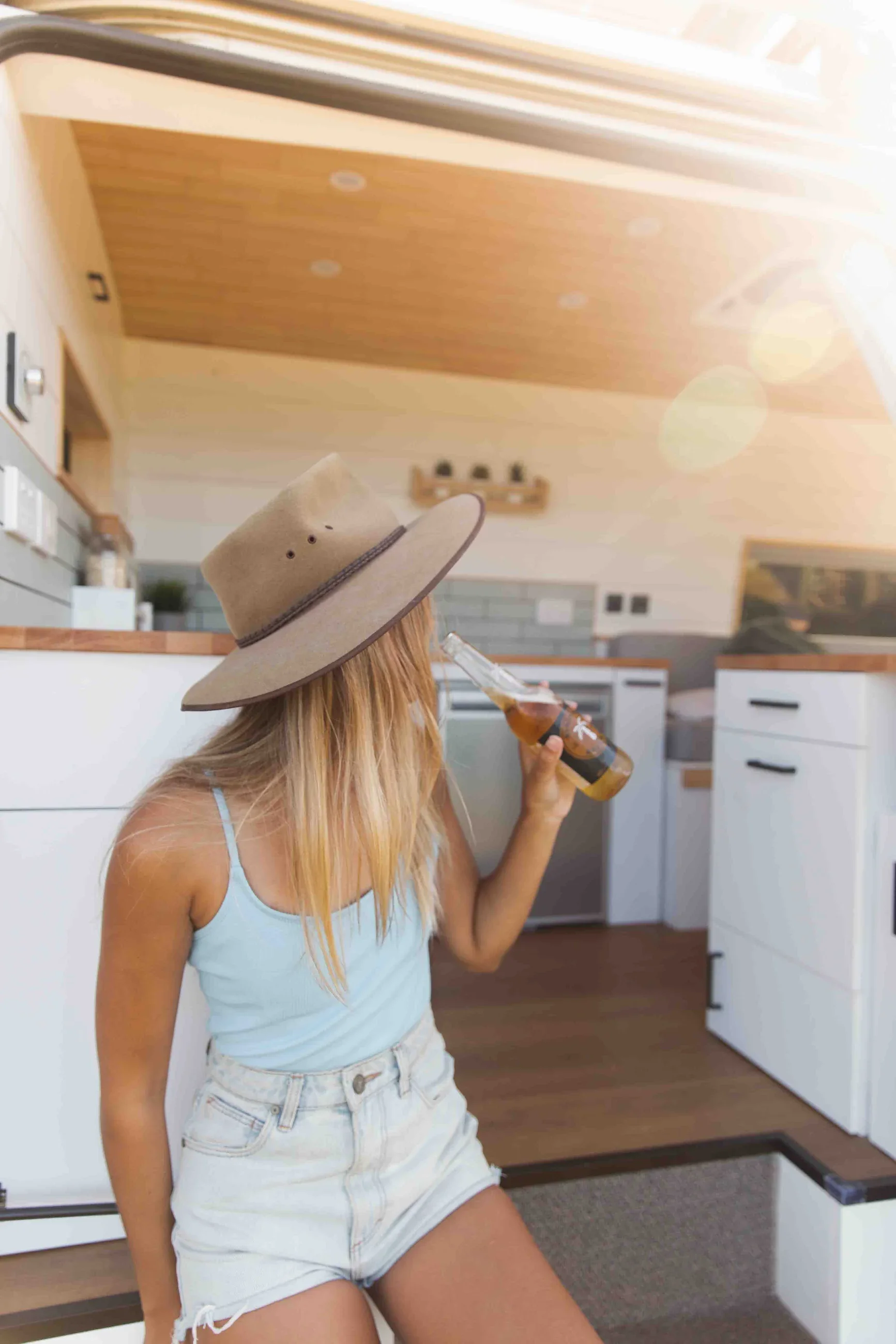 Person in a hat and light blue tank top drinks from a bottle, sitting in a bright, modern kitchen. Sunlight filters in from above.