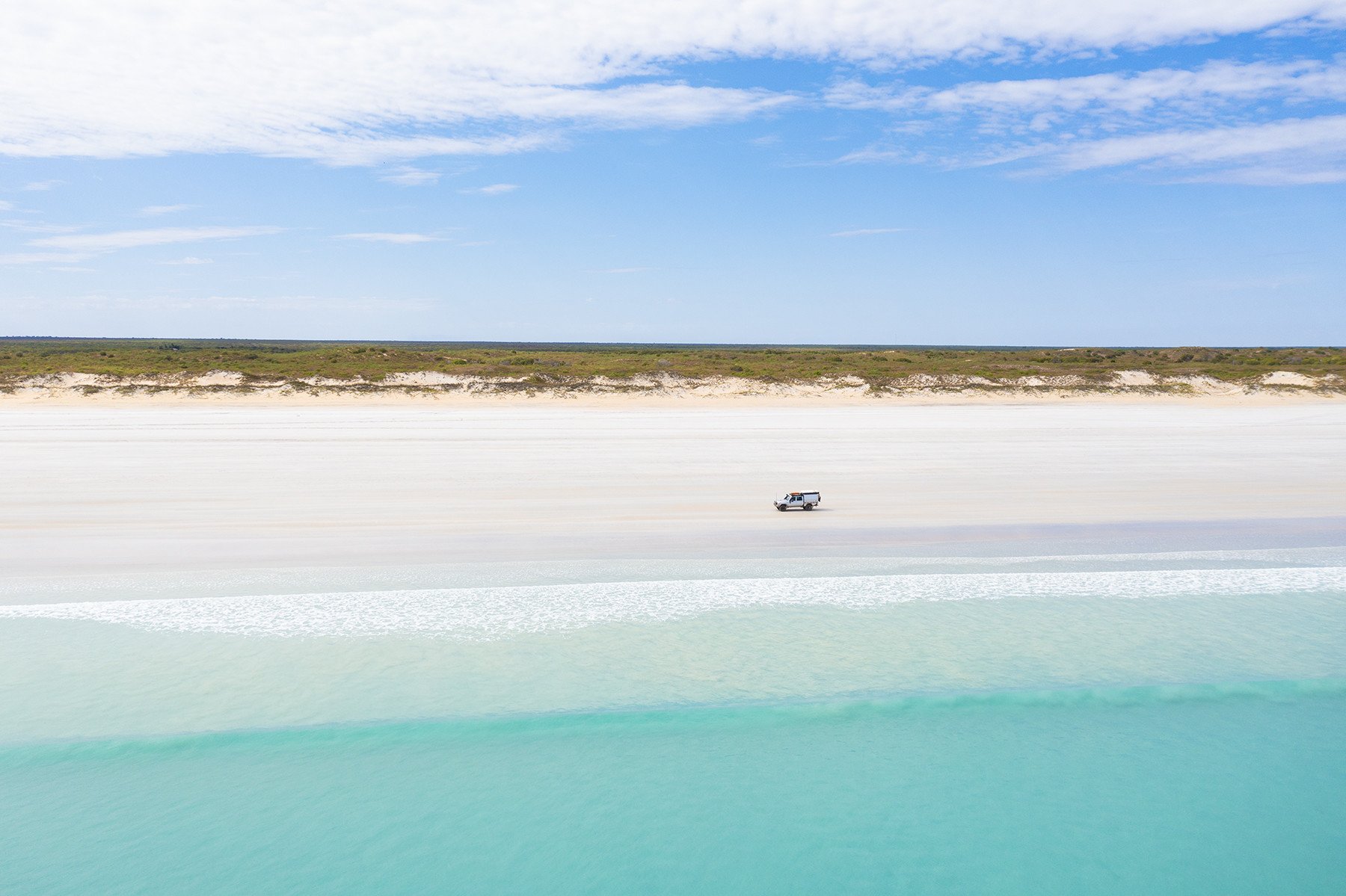Aerial view of turquoise ocean and wide white beach with a lone 4x4 parked near the shoreline, sand dunes and a blue sky with scattered clouds.