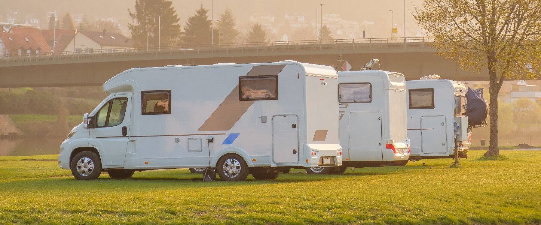 Autocaravana blanca estacionada en camping con toldo extendido, sillas y mesa fuera.