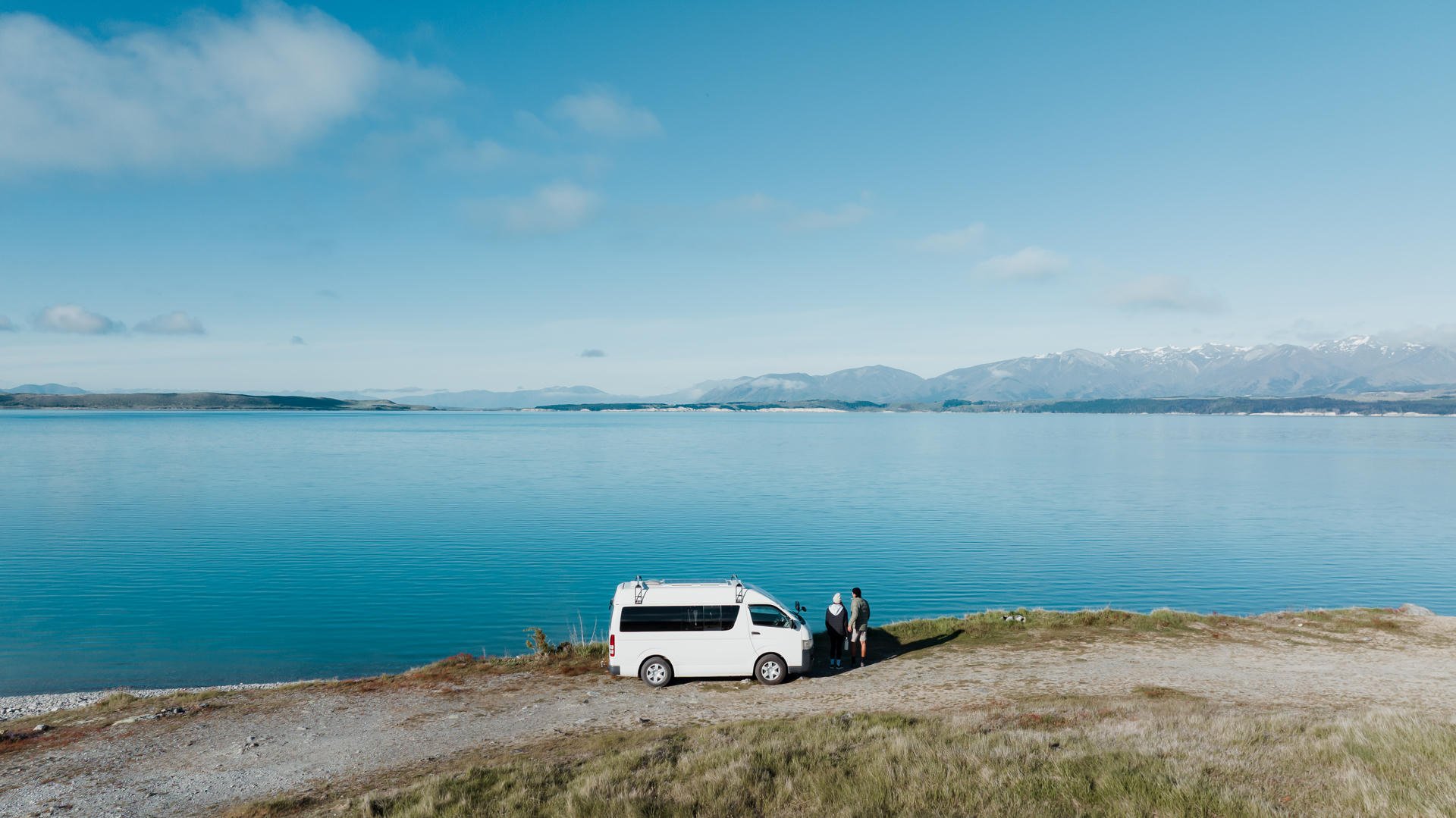 A white van parked by a tranquil lake with mountains in the background under a clear blue sky. Two people stand near the van.