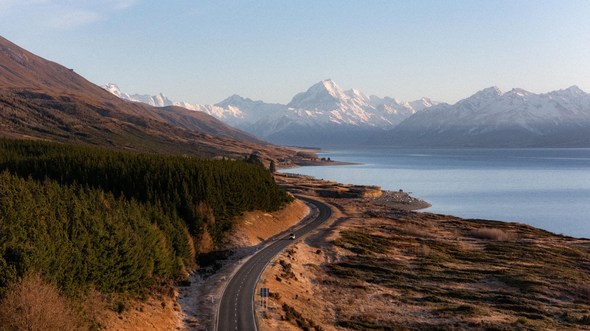 Winding road along a lake with a forest on one side, leading to snow-capped mountains under a clear blue sky.