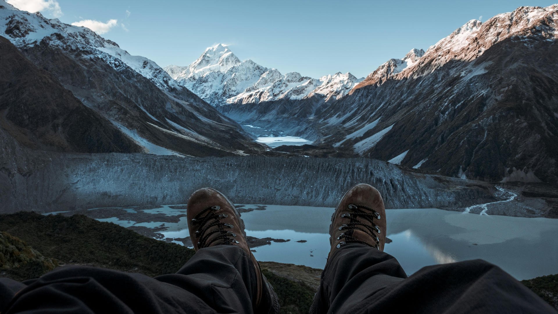 Hiker's boots in the foreground with a stunning view of snow-capped mountains and a serene lake under a clear blue sky.