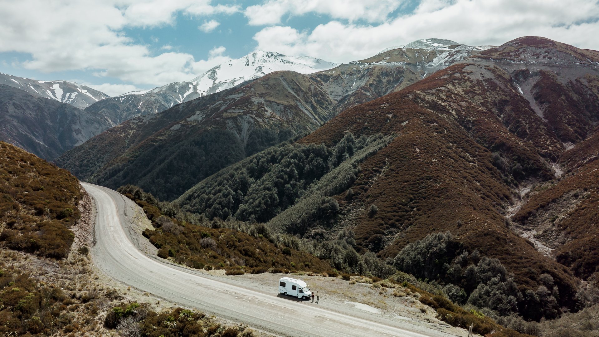 A white camper van parked on a winding mountain road, surrounded by rugged, snow-capped peaks and dense forests under a cloudy sky.