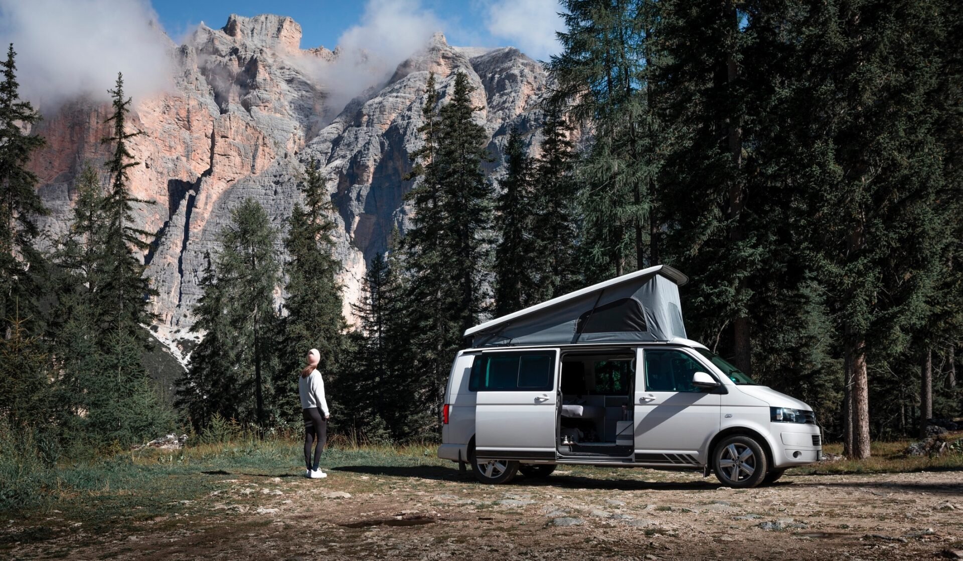 Eine Person steht neben einem Campervan mit offenem Dach, umgeben von hohen Bäumen und felsigen Bergen unter einem blauen Himmel.