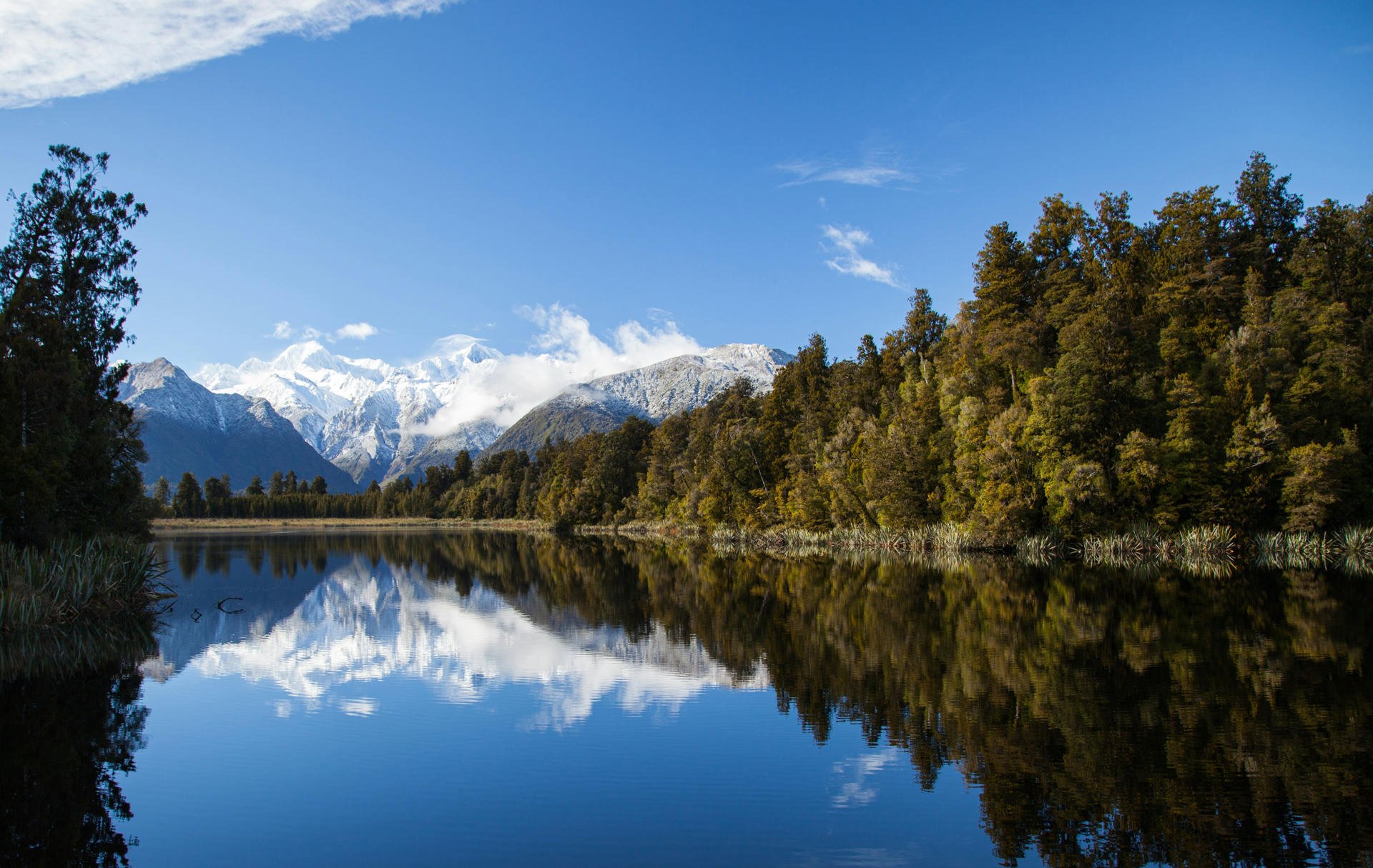 A serene lake reflects snow-capped mountains and lush trees under a clear blue sky, creating a tranquil and picturesque landscape.