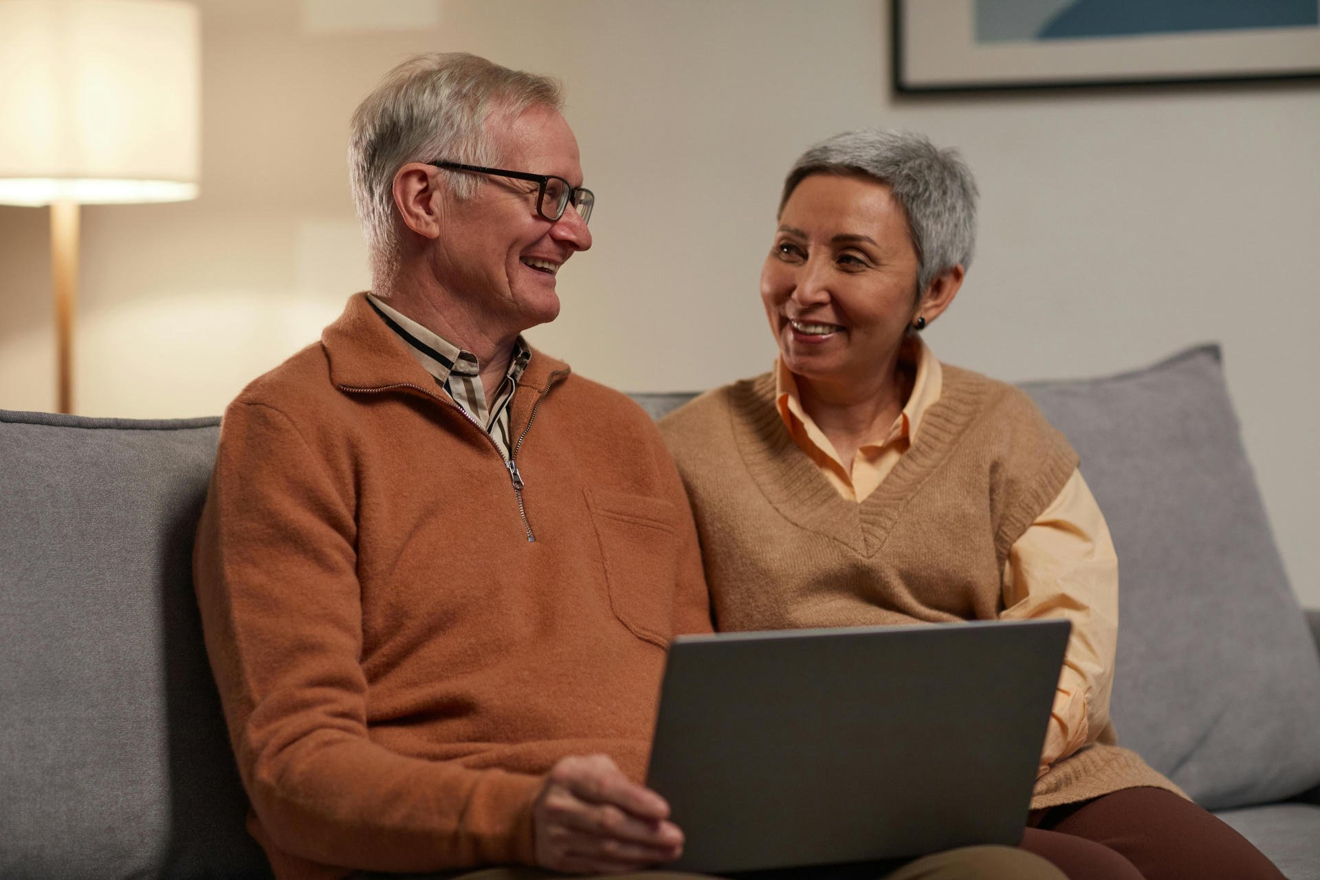 Elderly couple sitting on a couch, smiling at each other, with a laptop on their laps. They are wearing warm, casual clothing.
