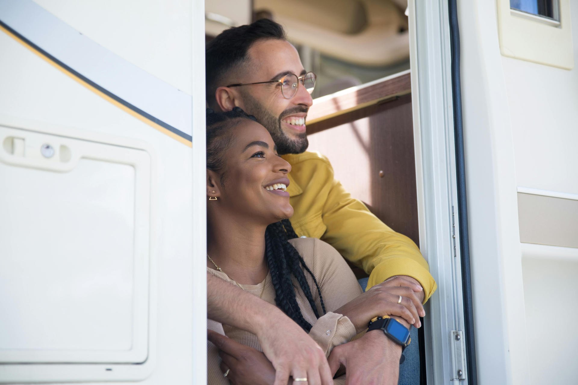 Couple smiling and embracing, sitting in the doorway of a camper van, enjoying a sunny day.