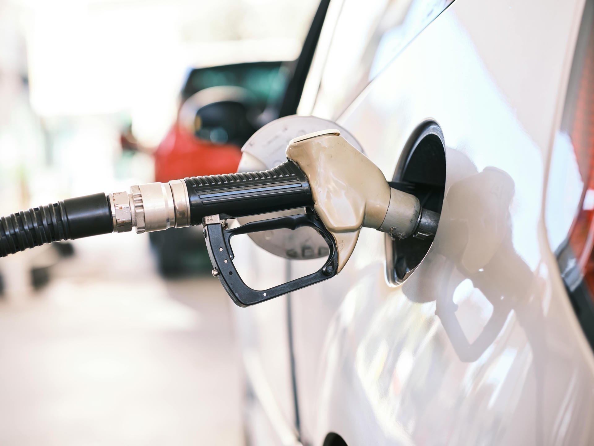 A fuel nozzle inserted into a car's gas tank at a filling station, with another car blurred in the background.