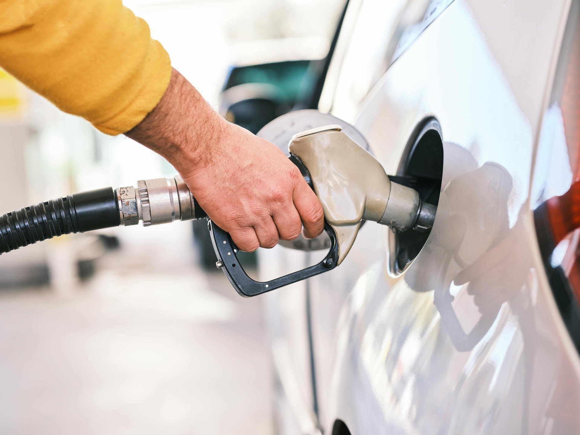 Person in a yellow sweater refueling a car at a gas station, holding a fuel nozzle inserted into the car's fuel tank.