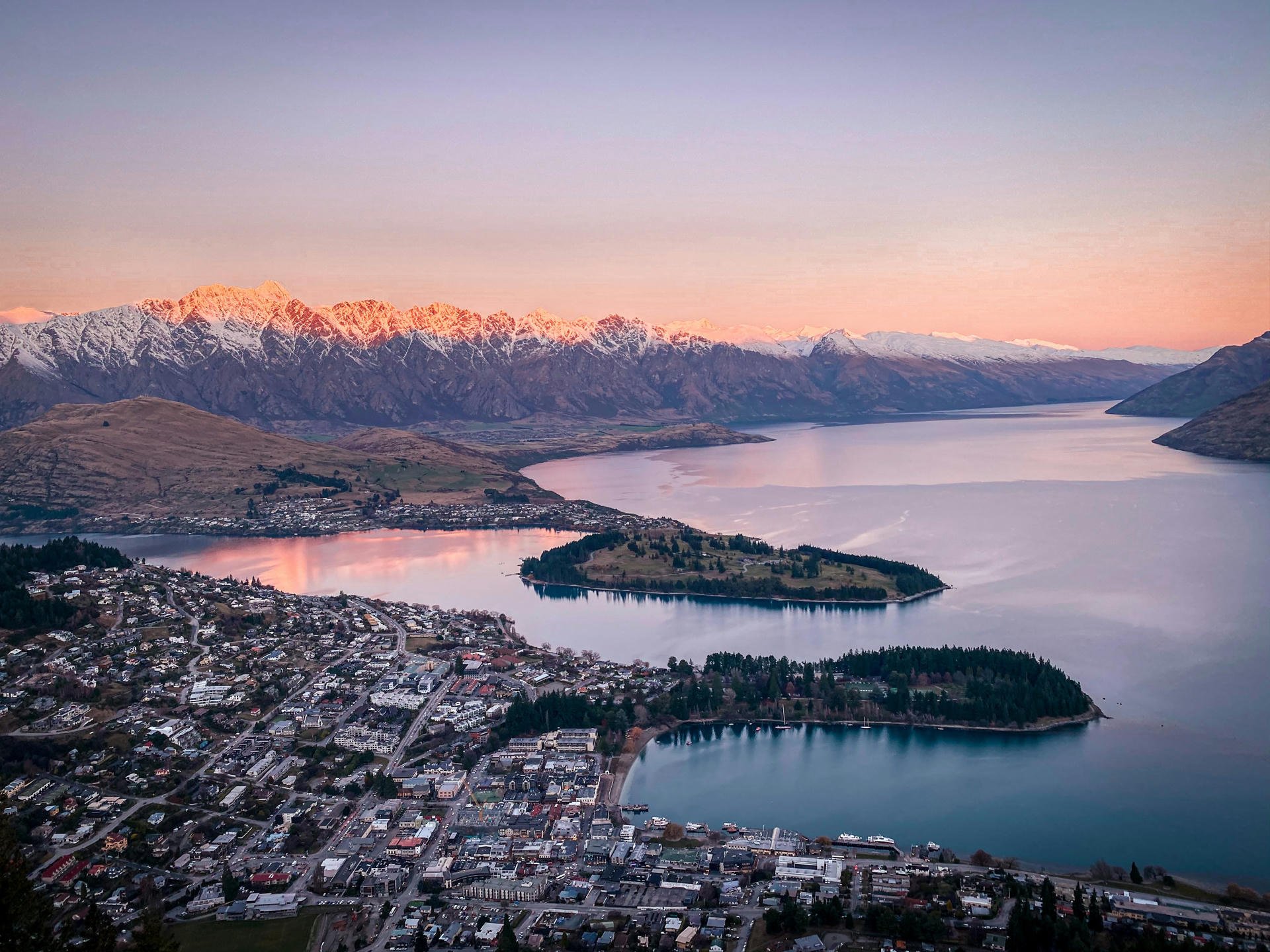 Aerial view of a town by a lake with snow-capped mountains in the background at sunset, reflecting hues of pink and purple in the water.
