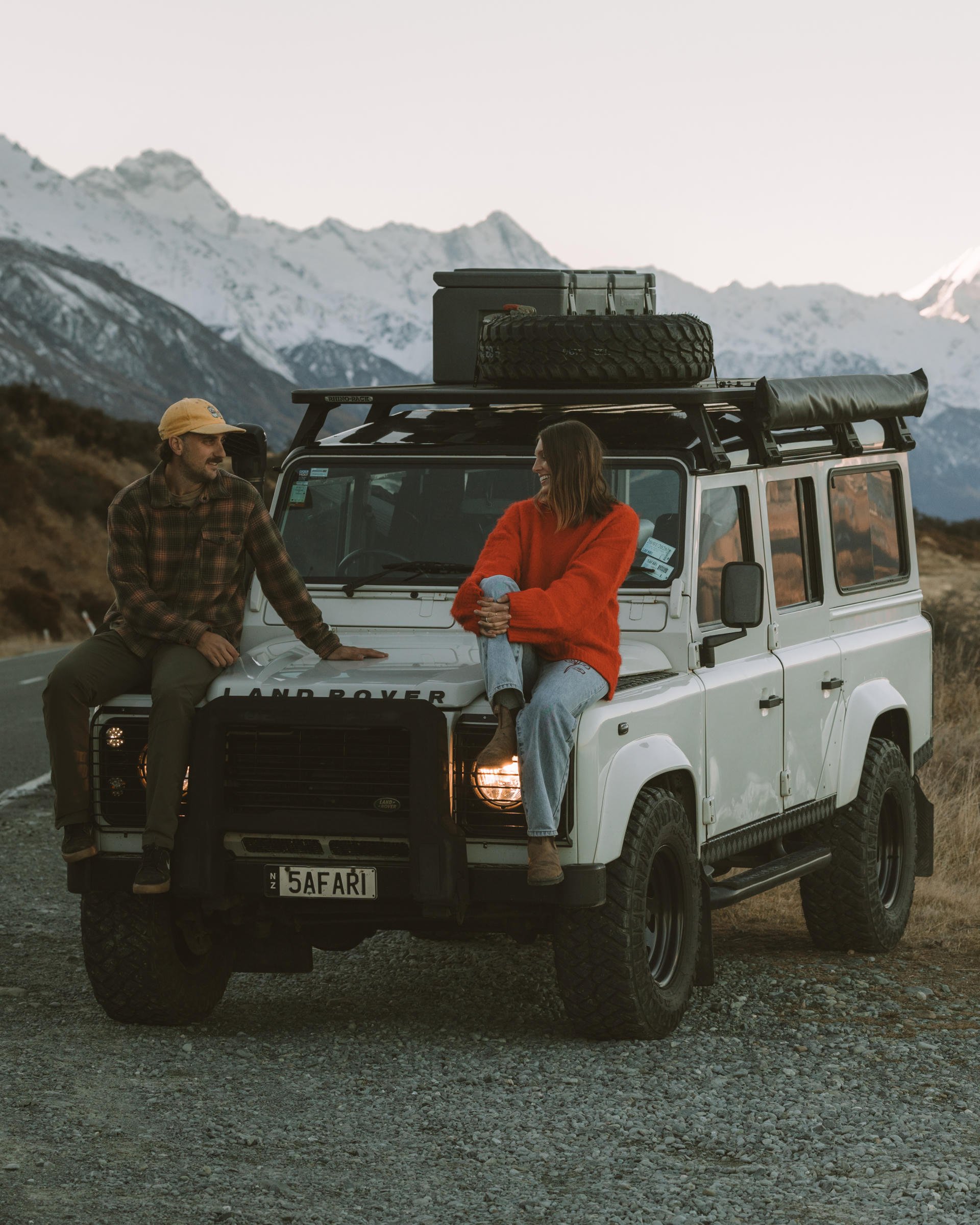 A man and woman sit on a white Land Rover parked on a mountain road, with snow-capped peaks in the background.