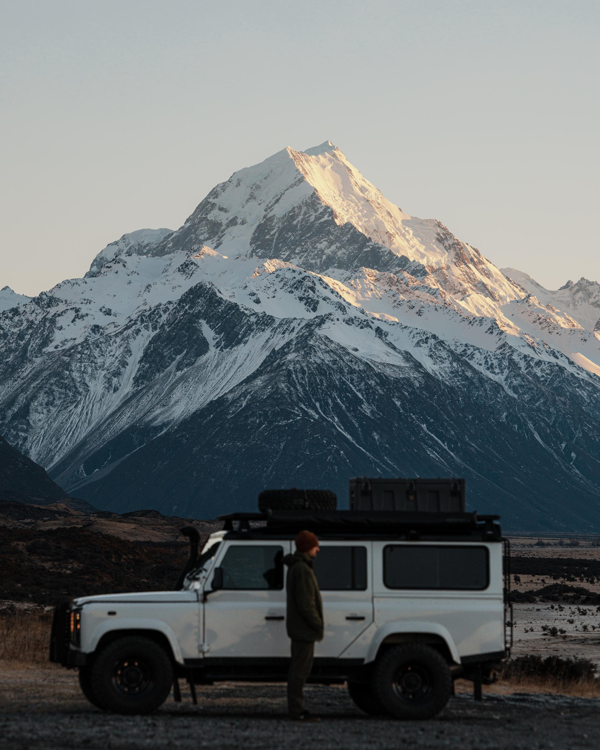 A person stands beside a white SUV with a roof rack, set against a backdrop of snow-capped mountains under a clear sky.
