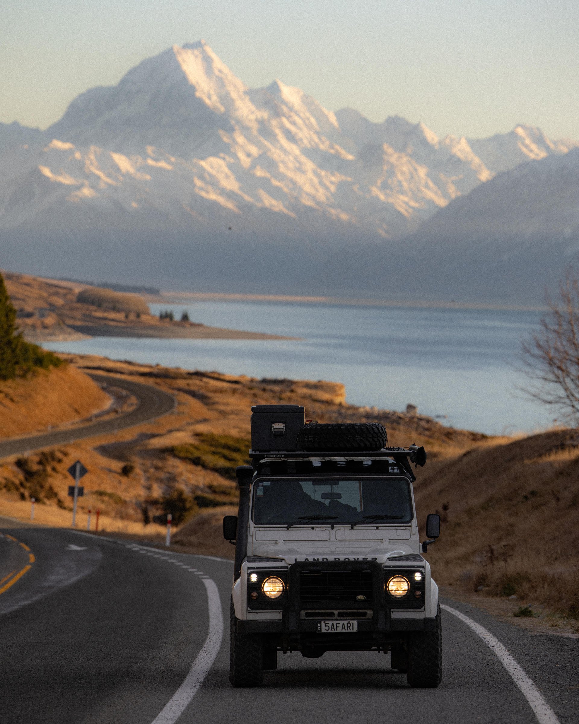 A rugged vehicle drives on a winding road with snow-capped mountains and a serene lake in the background under a clear sky.