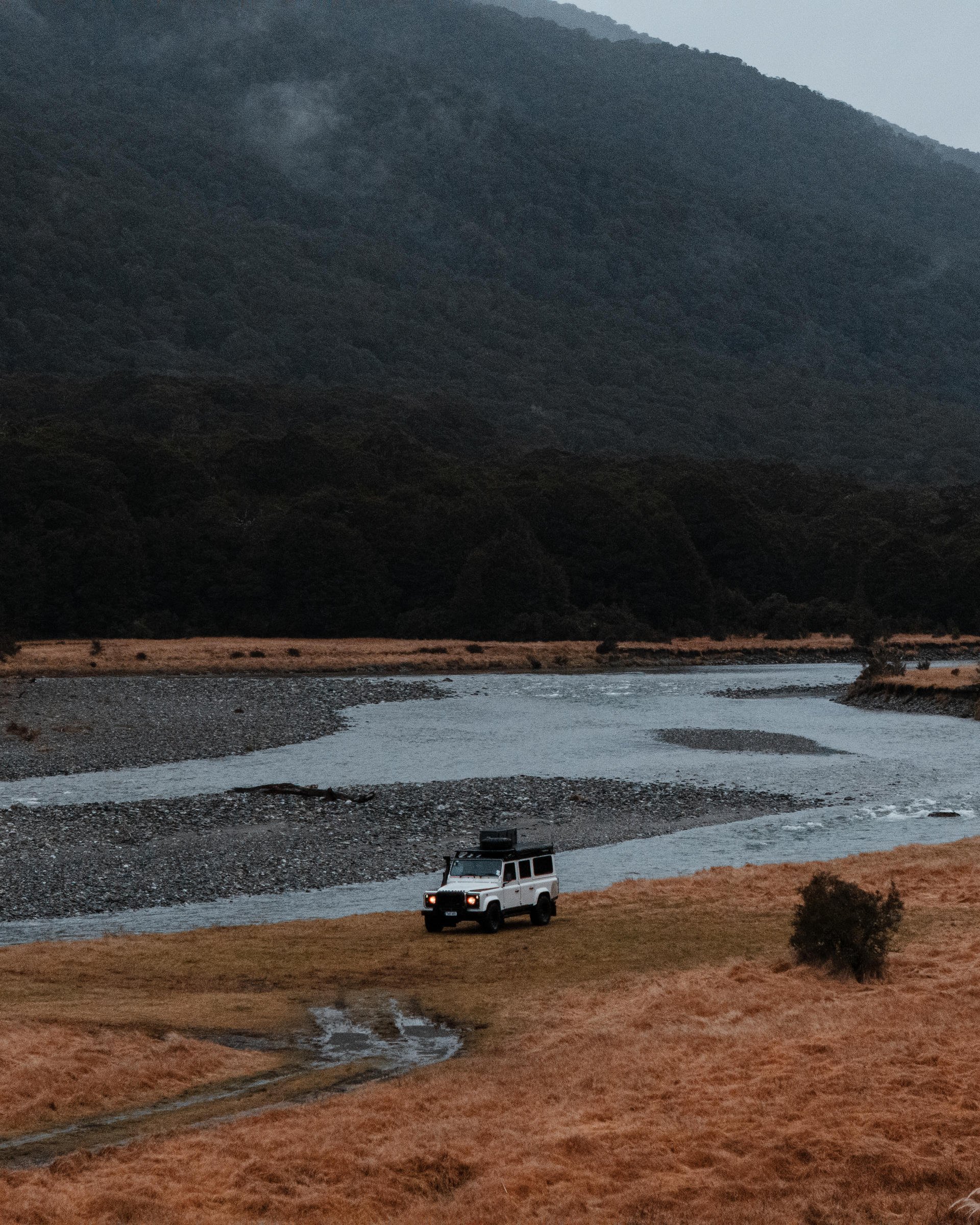 A white off-road vehicle drives along a winding river in a mountainous landscape with overcast skies and grassy terrain.