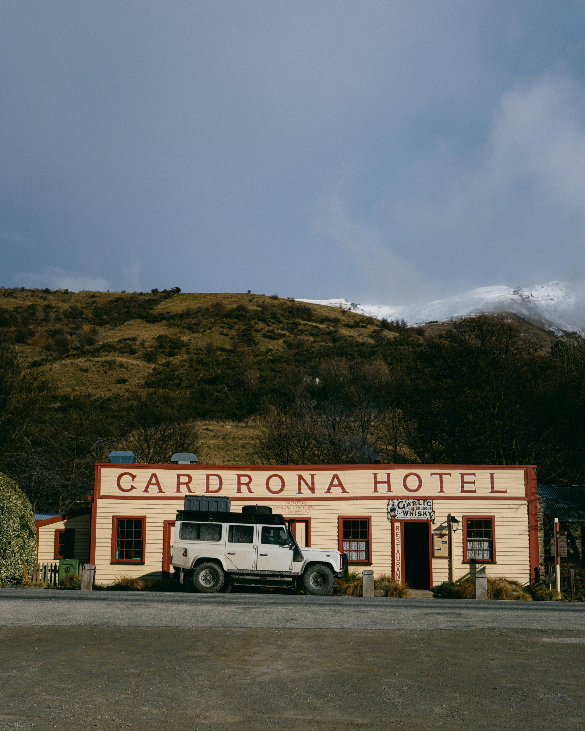 A vintage white vehicle is parked in front of the historic Cardrona Hotel, set against a backdrop of hills and a partly cloudy sky.