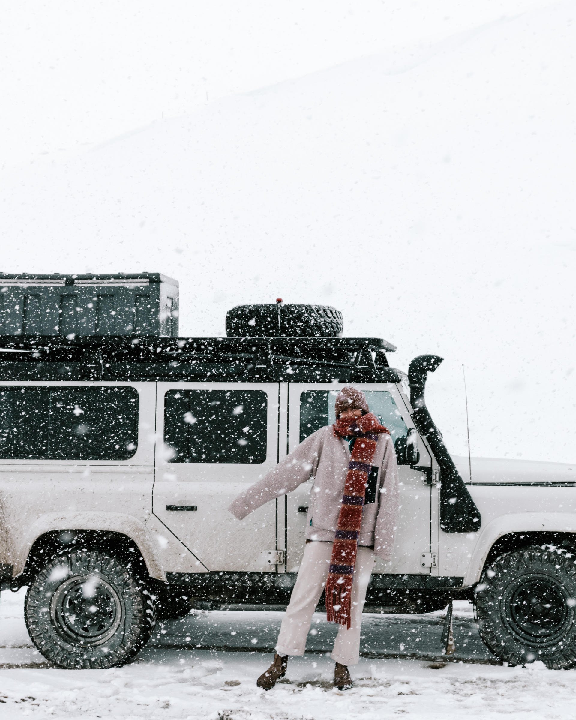 Person in winter attire stands by a snow-covered SUV in a snowy landscape, wearing a red scarf and beanie.