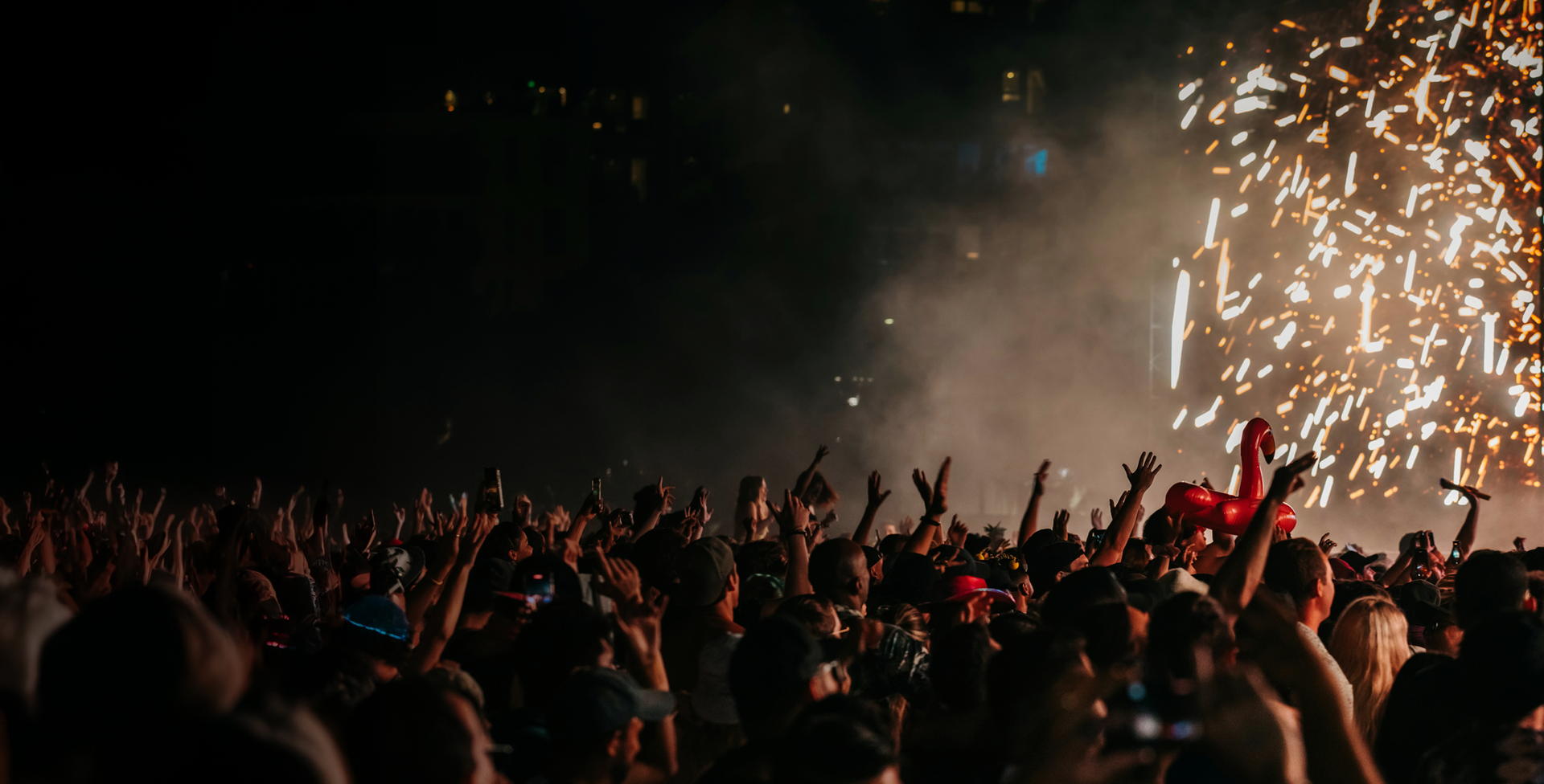 A crowd at a concert raising their hands, with bright fireworks illuminating the night sky in the background.