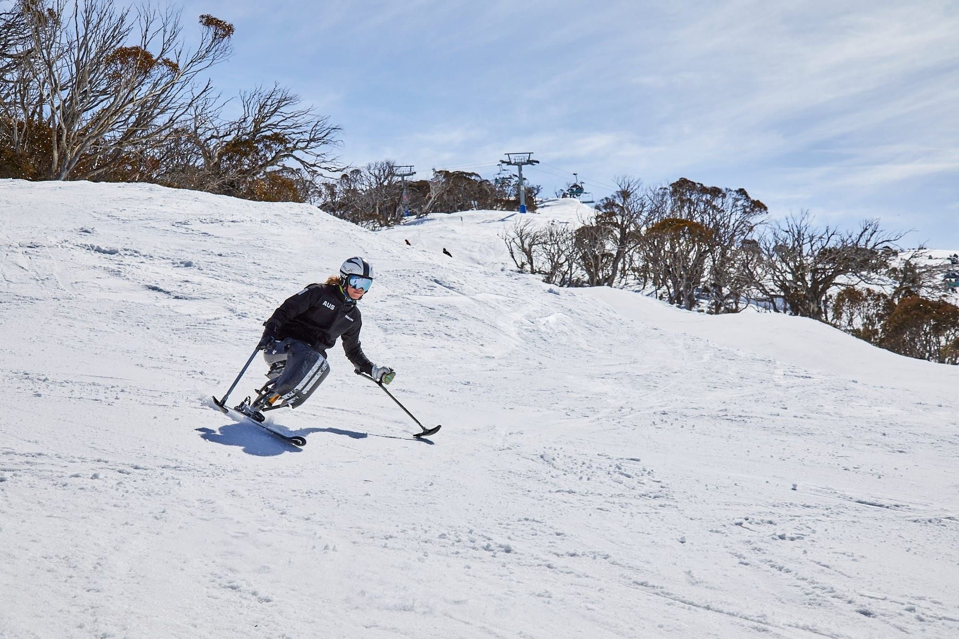 Adaptive skier on a monoski using outriggers carving down a snowy slope with trees and a chairlift in the background.