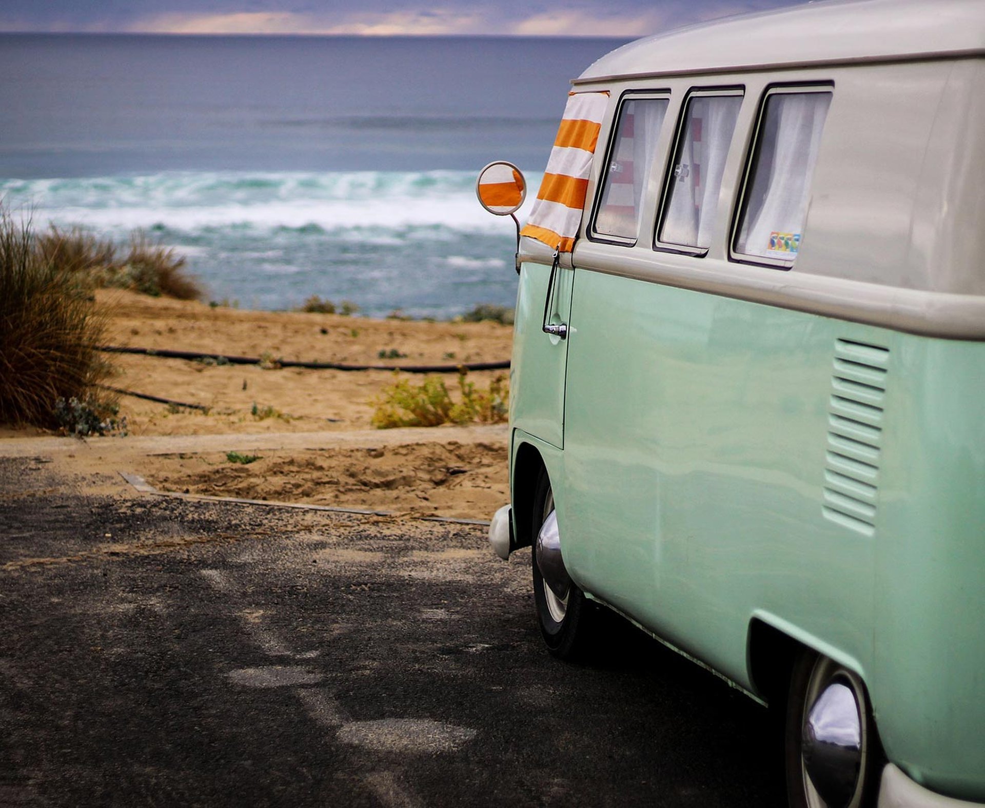 VW Camper in hellblau steht an der Küste am Strand, orange-weiße Handtücher hängen als Sonnenschutz in den Fenstern