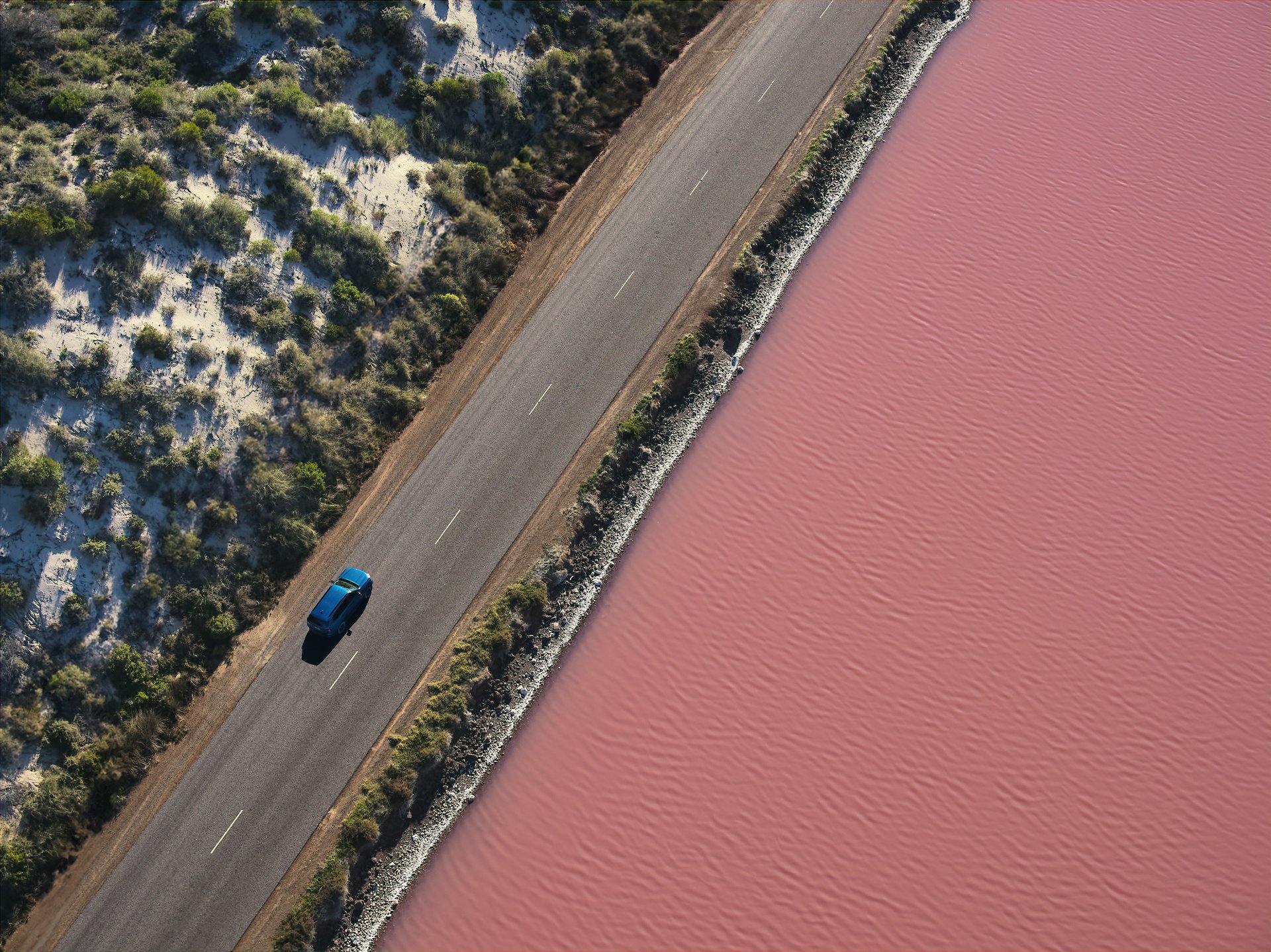 Aerial view of a lone blue car driving along a narrow road between scrubby sand and a pink salt lake.