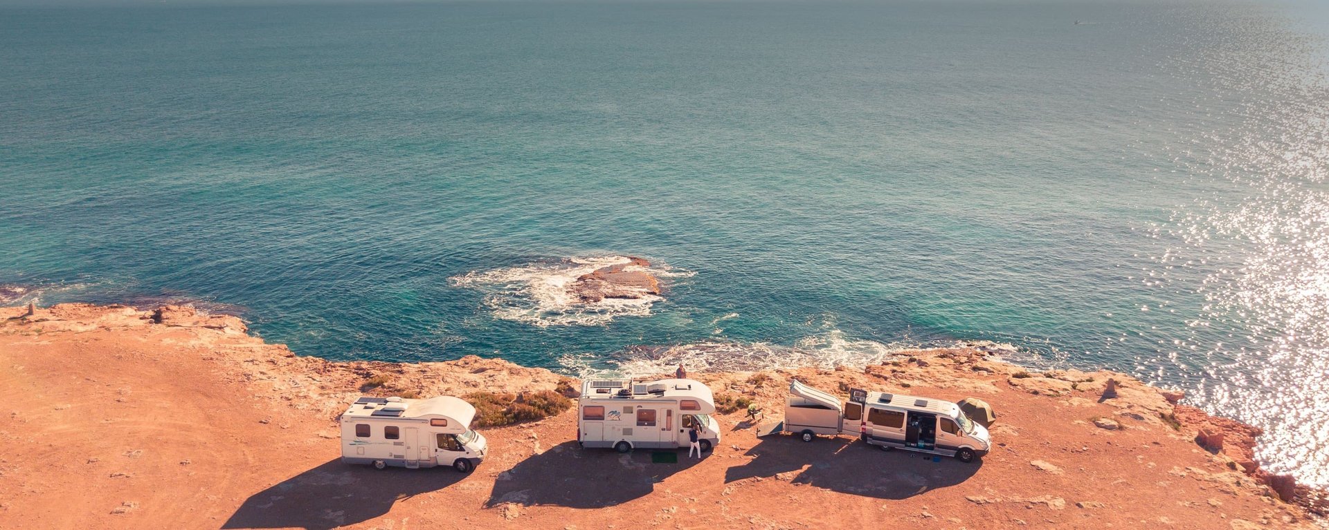 Three RVs parked on a rocky coastal cliff overlooking a vast, sparkling blue ocean under a clear sky.