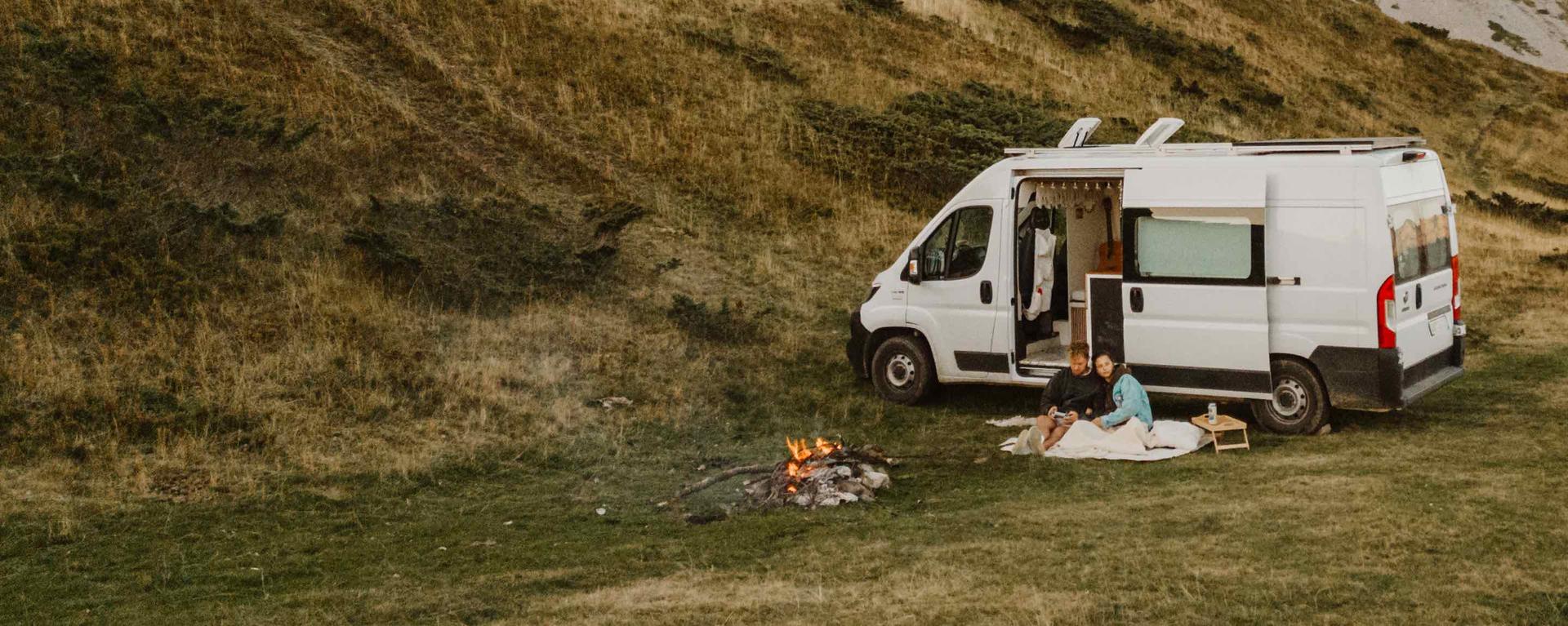 A white camper van parked on grass beside a campfire, with two people sitting on a blanket, surrounded by hills.