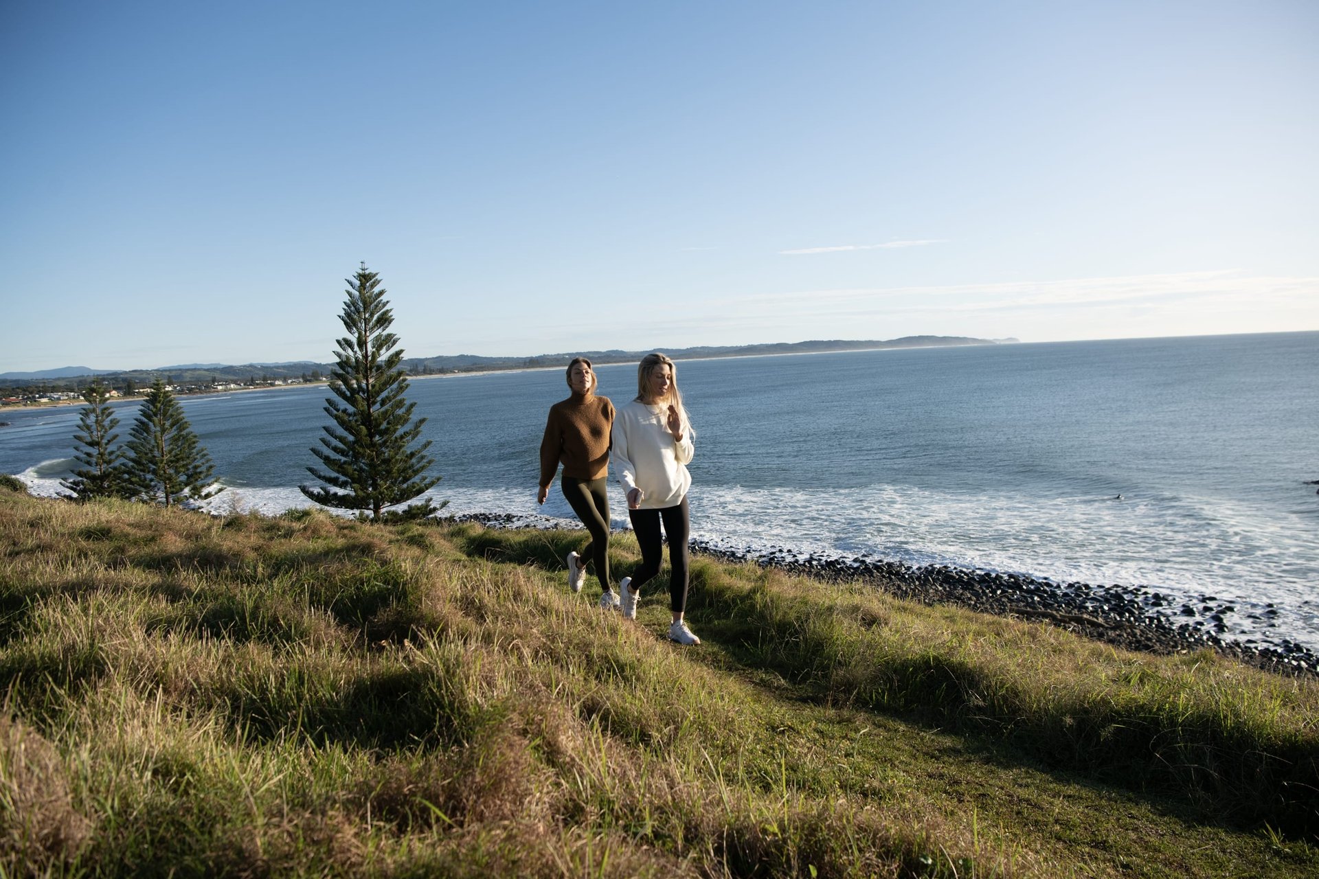 Two people walking along a grassy coastal bluff with pine trees and a calm ocean under a clear sky.