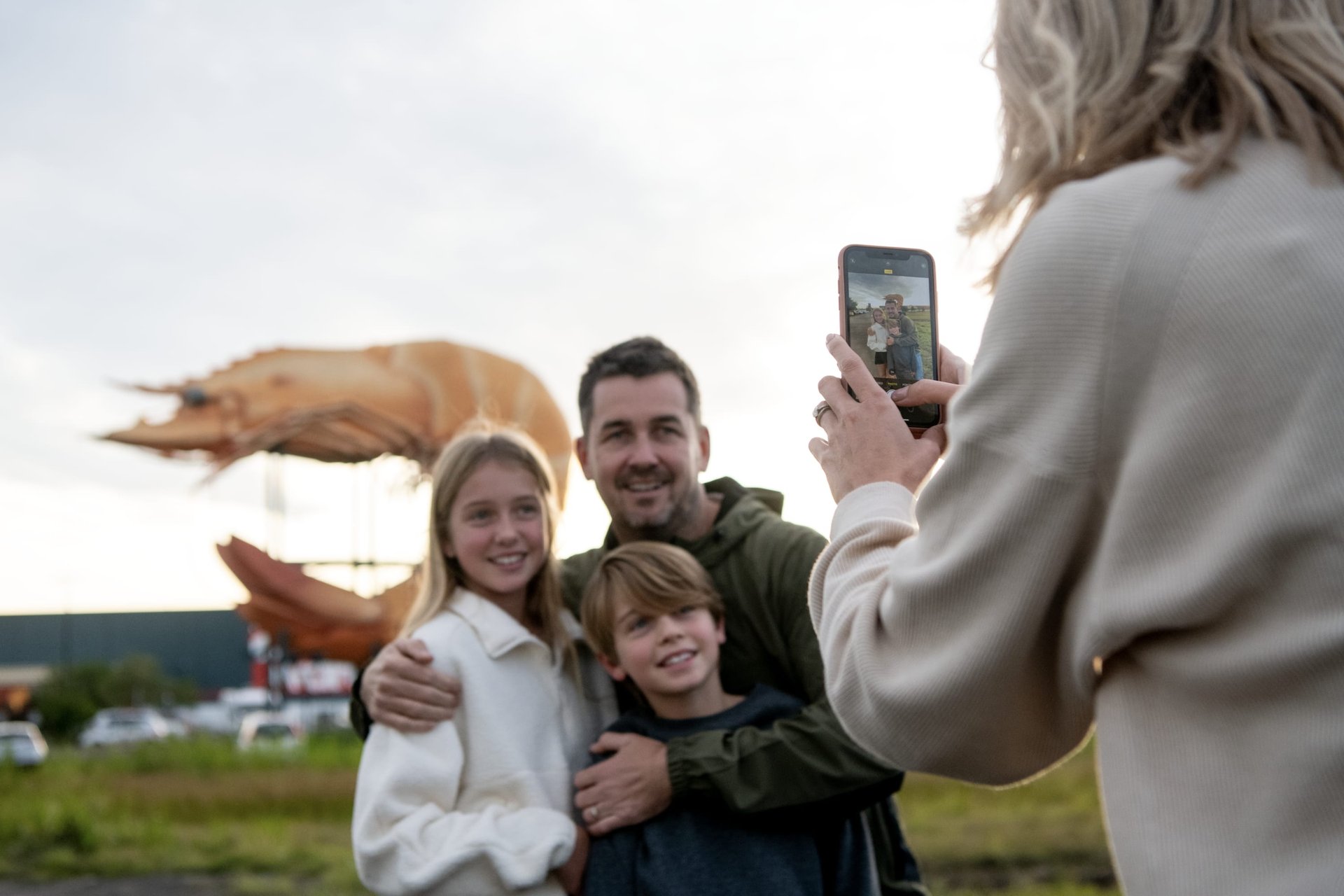 Person photographing a smiling father and two children posing outdoors in front of a giant shrimp sculpture.