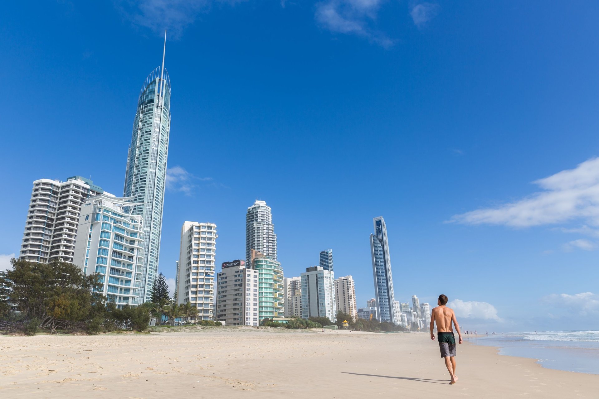 A person walks along a sandy beach with a backdrop of tall, modern skyscrapers under a clear blue sky.