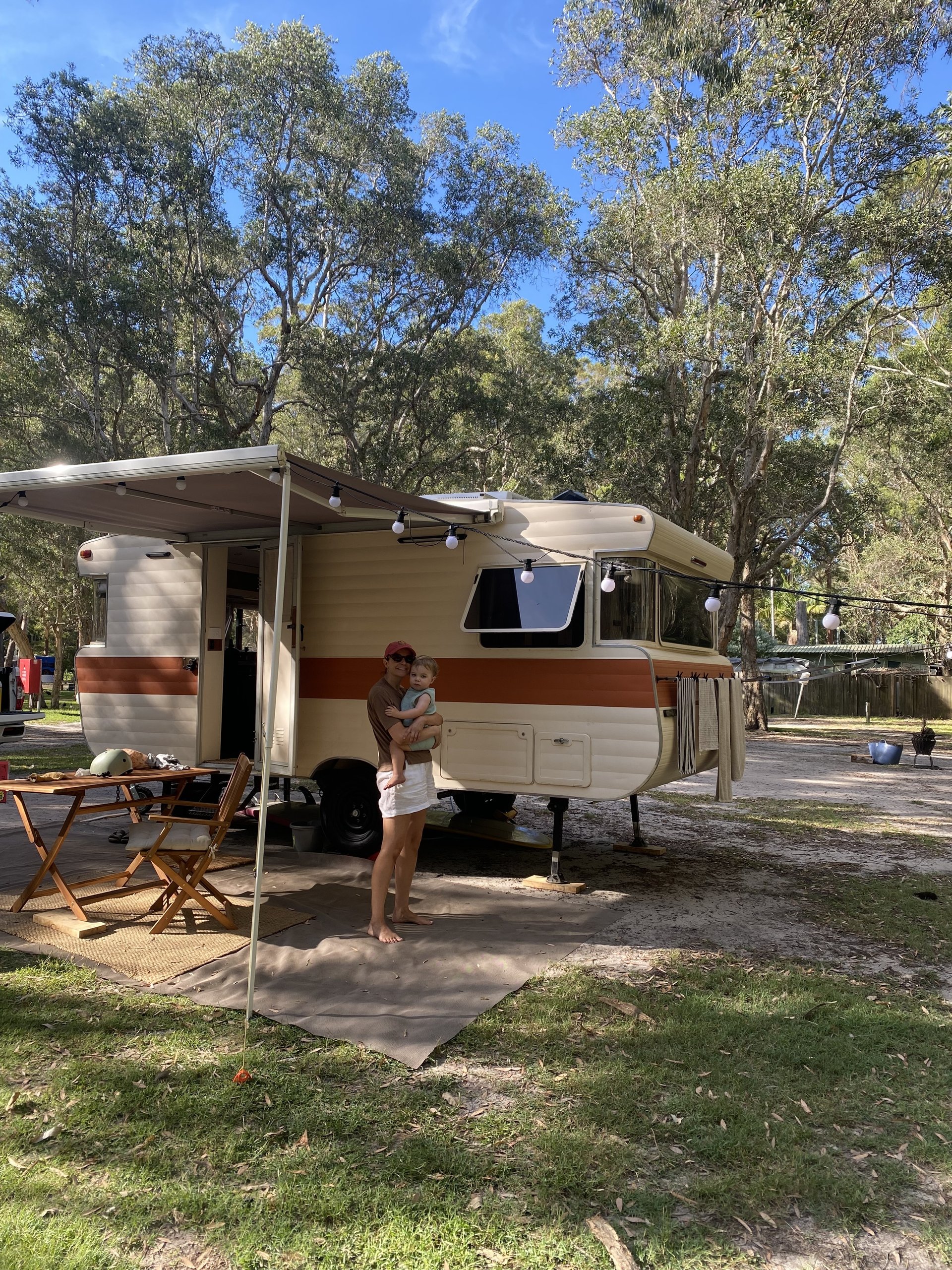 A person holding a baby stands outside a vintage camper with an awning and string lights, surrounded by trees and outdoor furniture.