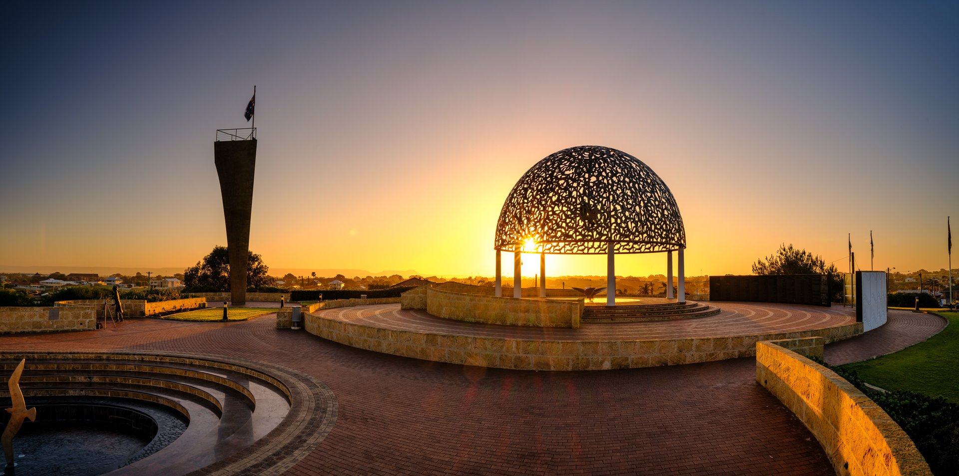Sunset behind an ornate lattice metal dome pavilion with circular stone amphitheater and nearby flag-topped tower.