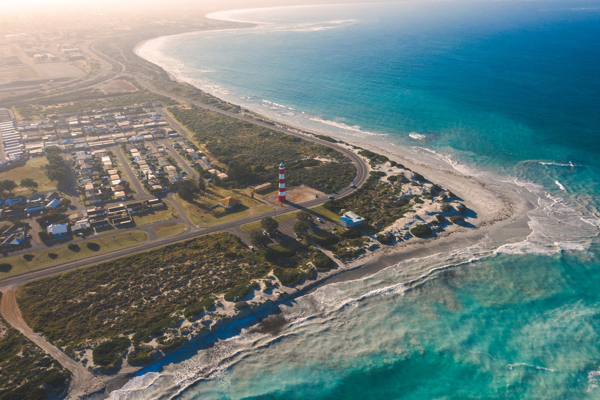 Aerial view of coastline with turquoise sea, sandy beach, curved road, residential neighborhood, and red-and-white striped lighthouse.