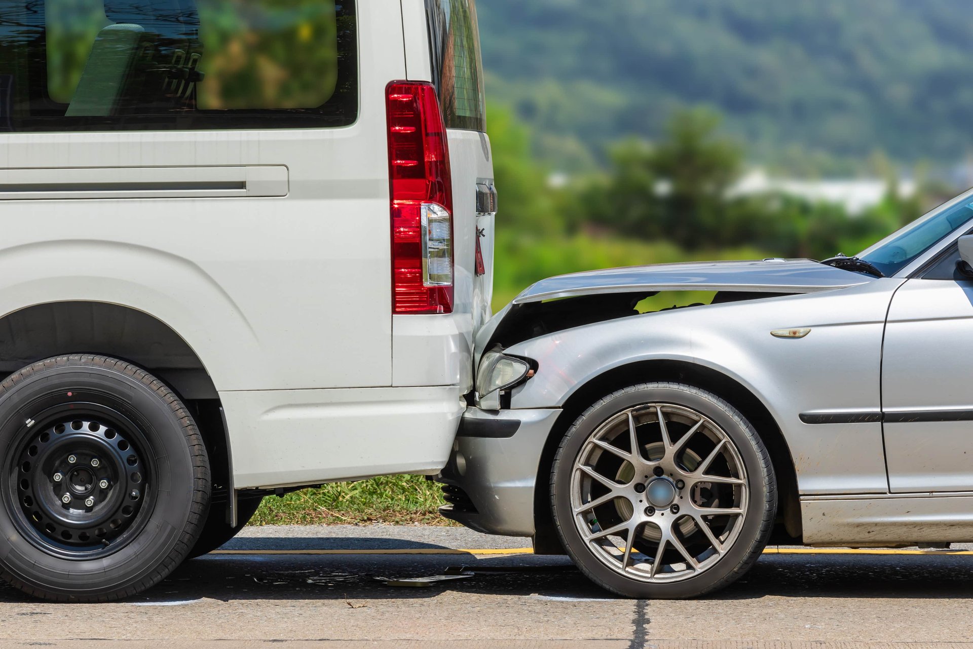 A silver car with a damaged front bumper is rear-ended into a white van on a road, with greenery in the background.