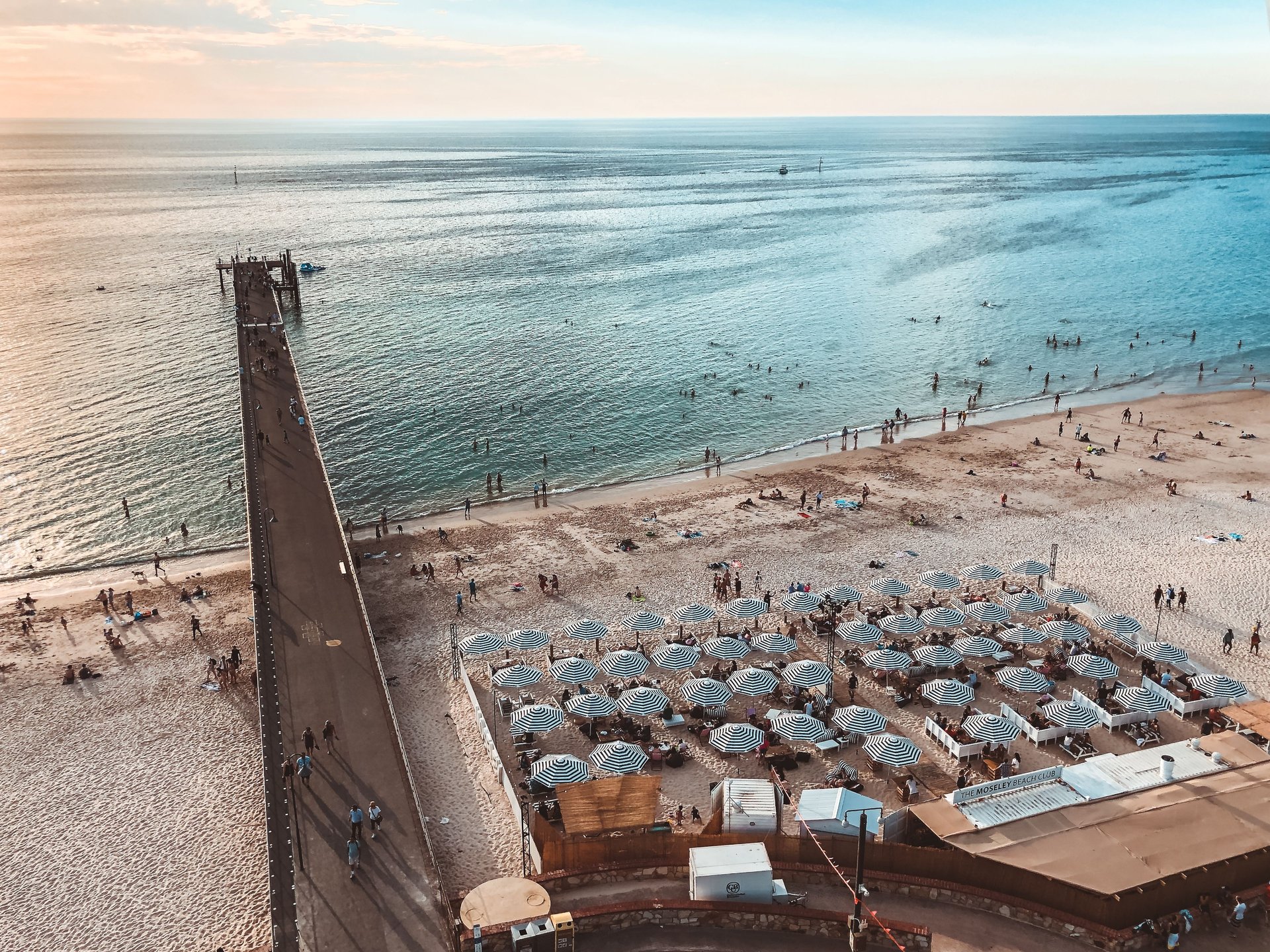 Aerial view of a beach with sun umbrellas, people swimming, and a long pier extending into the calm sea under a clear sky.