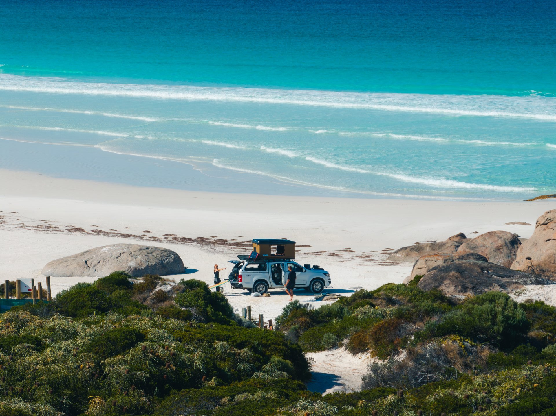 A vehicle with rooftop tent parked on a beach, surrounded by 2 people