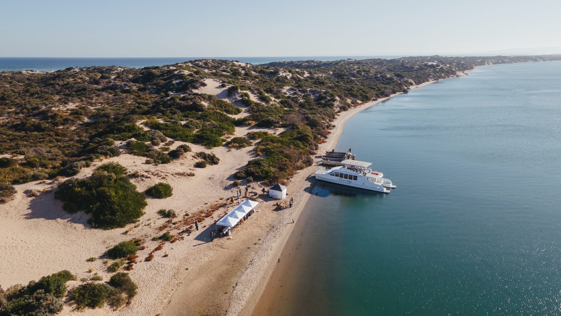 Aerial view of a sandy beach with white tents, lush greenery, and a boat docked by the shoreline, surrounded by calm, clear blue water.
