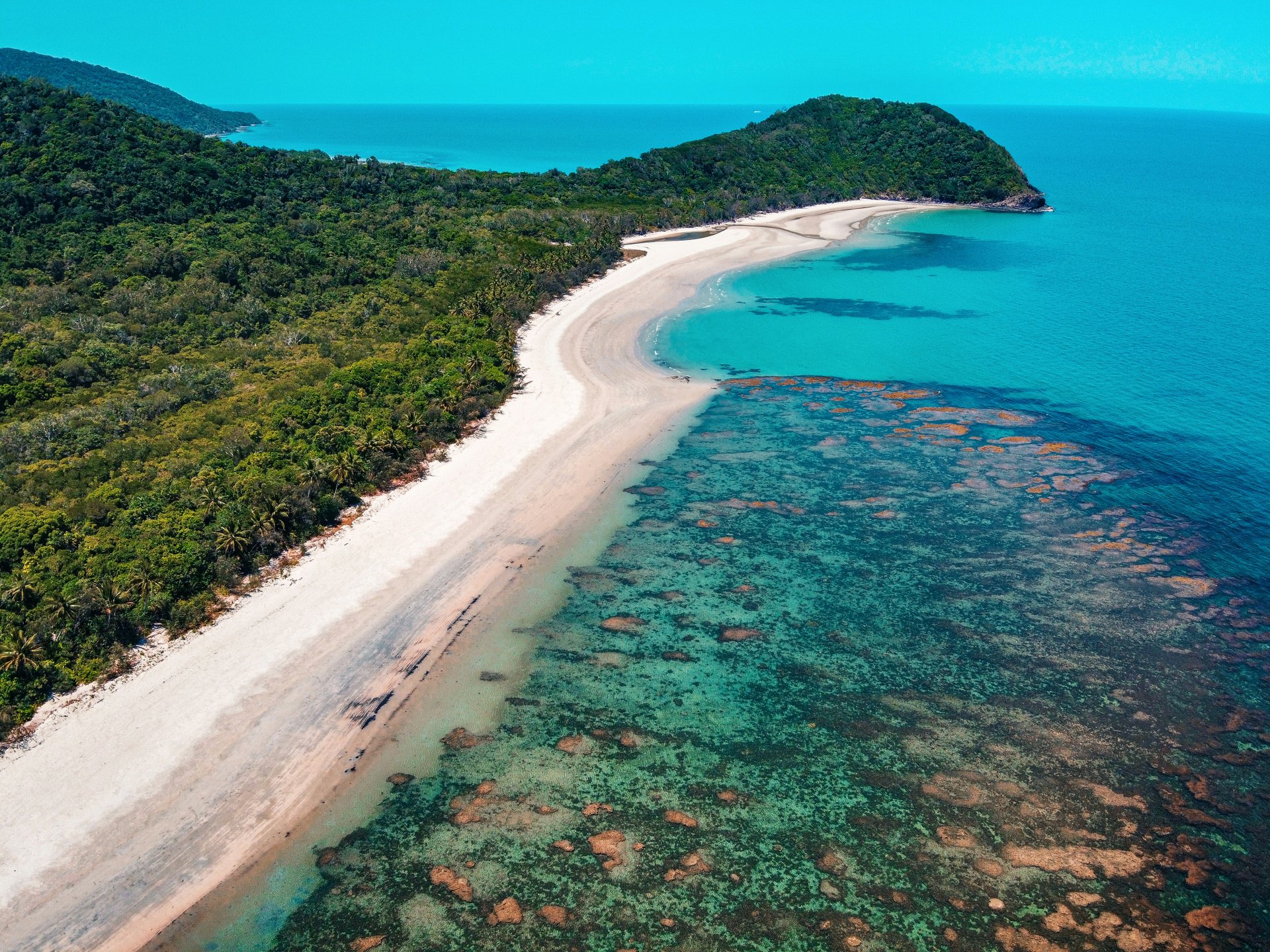 Aerial view of a tropical beach with white sand, lush green forest, and clear turquoise water, featuring visible coral reefs.