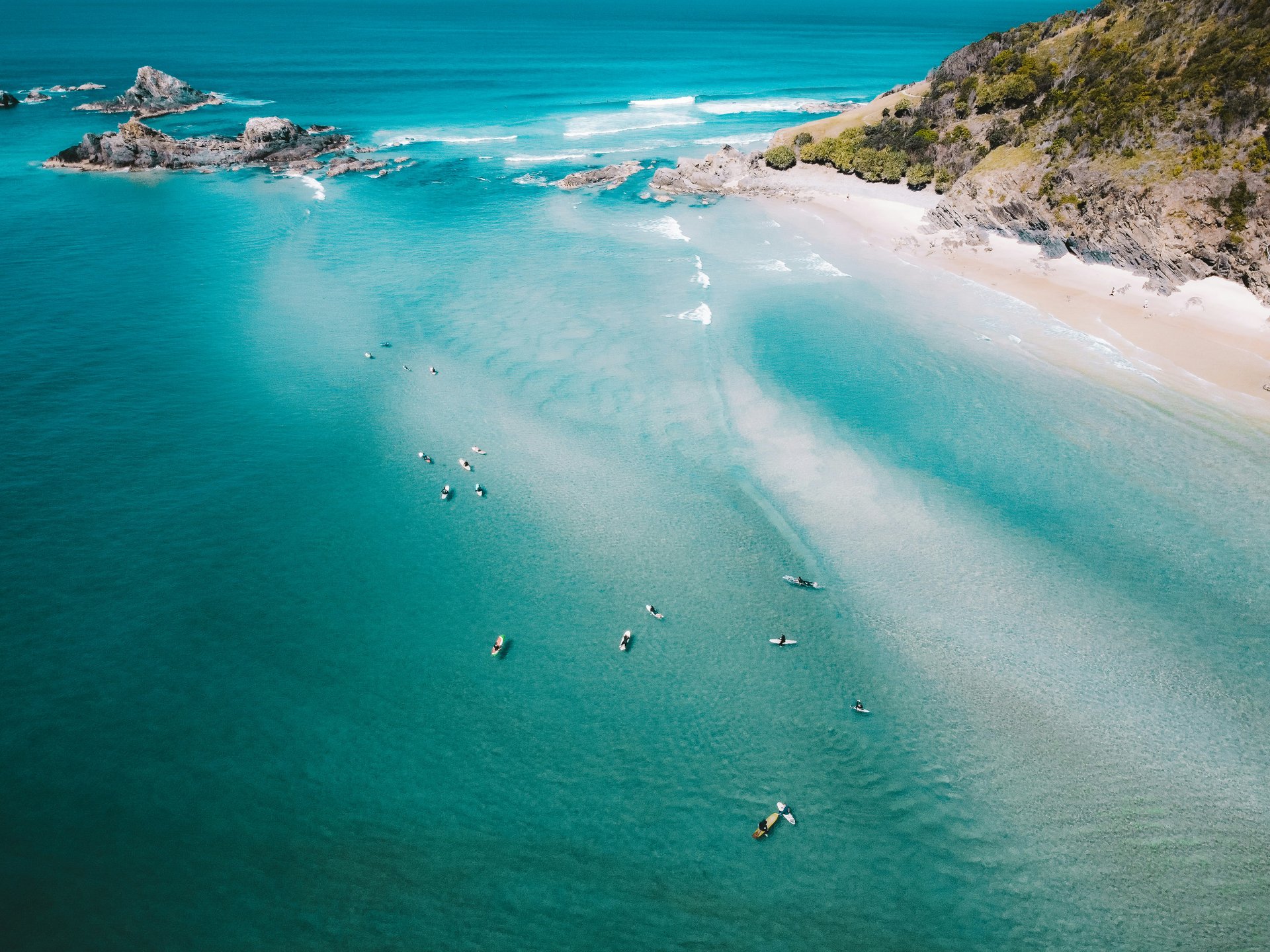 Aerial view of a turquoise beach with rocky outcrops, white sand, and people kayaking, surrounded by green hills.