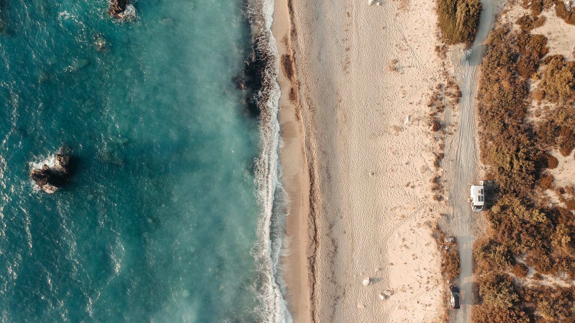 Luftaufnahme eines Sandstrandes mit türkisblauen Wellen, die an die Küste schlagen; ein weißes Fahrzeug, das auf einem Feldweg neben dem Strand geparkt ist.