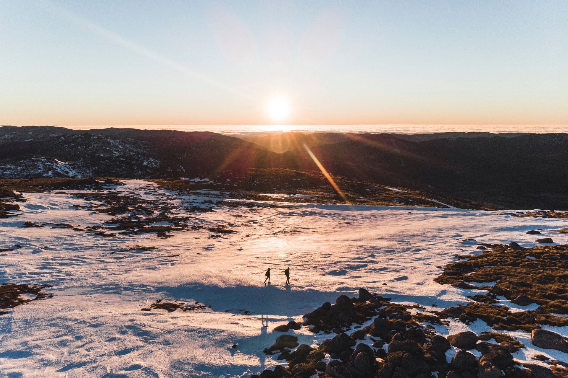 Two people walk on a snowy mountain landscape at sunrise, casting long shadows. The sun is low on the horizon, illuminating the scene.