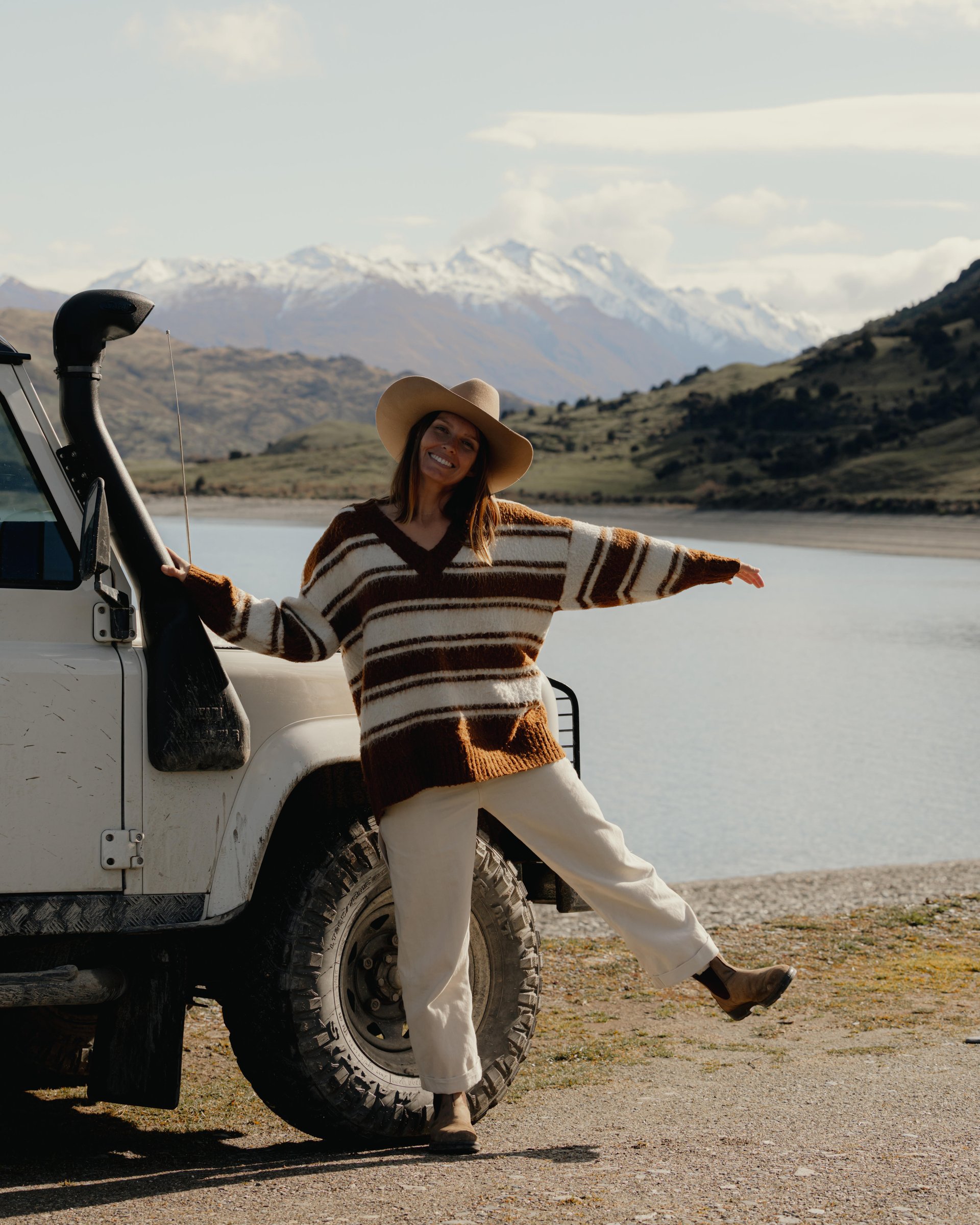 Woman in a striped sweater and hat joyfully poses by a white SUV near a serene lake, with snow-capped mountains in the background.