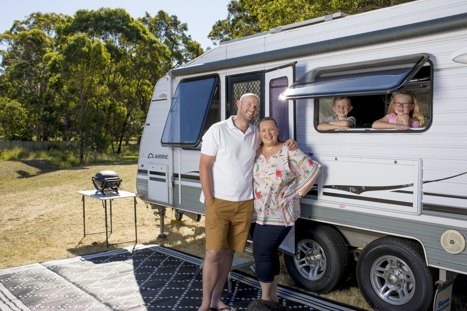A smiling couple stands beside a caravan in a sunny park, with two children peeking out of a window. A small grill is on a table nearby.